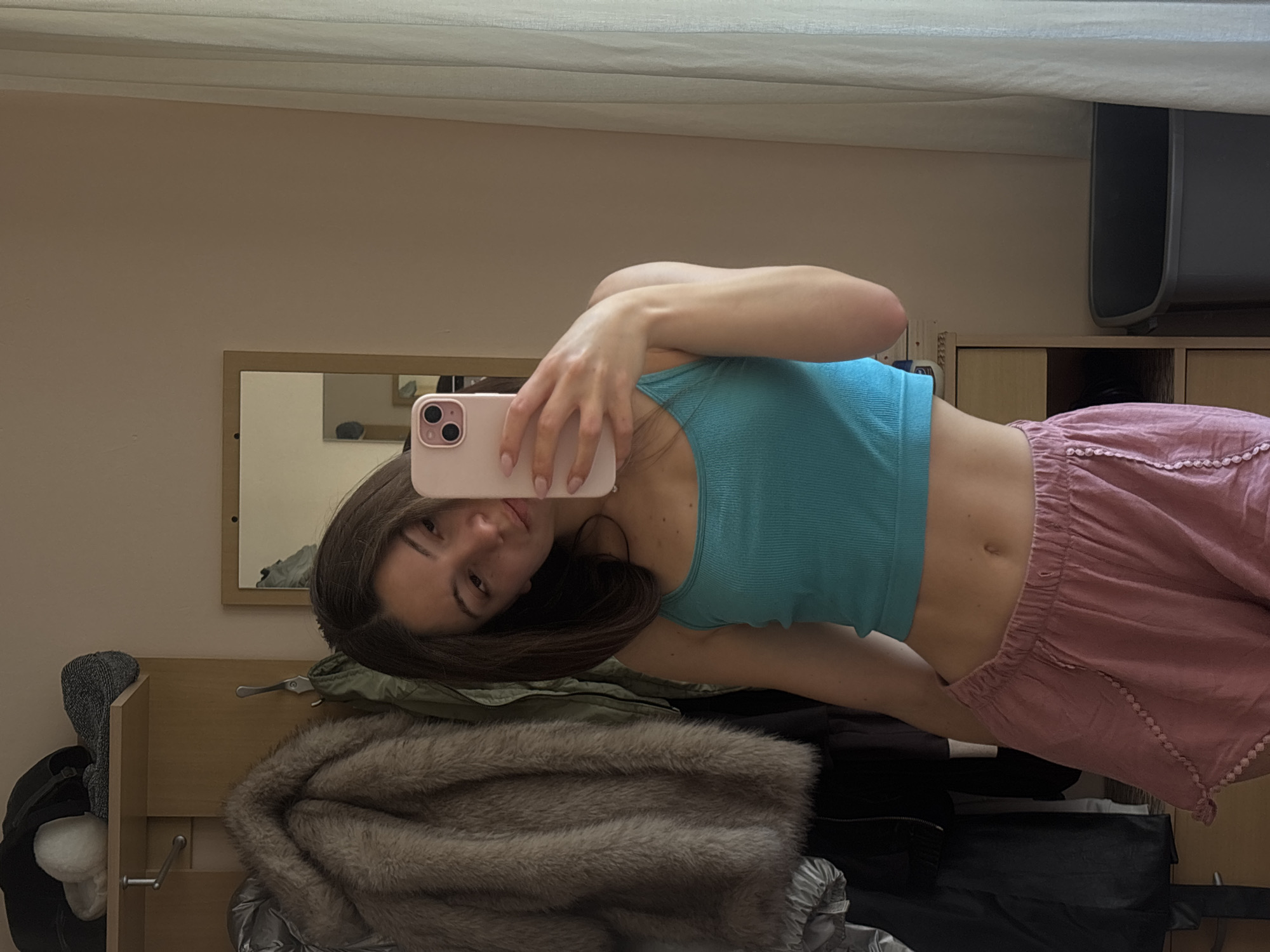 A young woman is taking a mirror selfie indoors. She is wearing a teal ribbed crop top and pink drawstring shorts. Her hair is dark brown and she is holding a pink phone in her right hand to take the picture. Behind her, a closet door is open, revealing several coats and jackets hanging on hooks. A mirror reflects a small portion of the room, showing what appears to be a window with sheer curtains and a pale beige wall. The overall setting suggests a bedroom or dressing area. There is no indication of the specific location within Varna, Bulgaria, as the image only shows an interior scene.