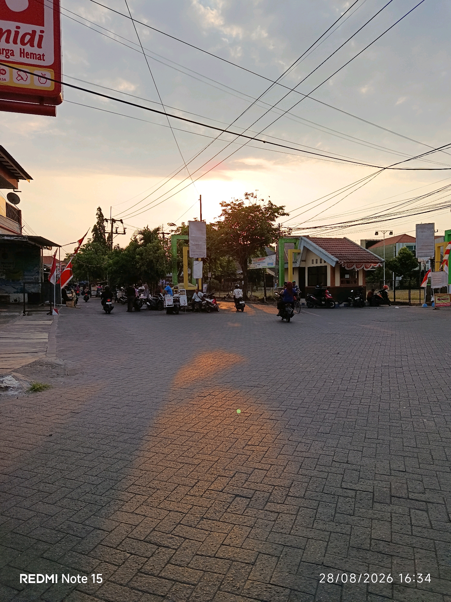 The image depicts a street scene in Keboansikep, Indonesia, during what appears to be sunset. The sky is partly cloudy with the sun visible low on the horizon, casting a warm glow. Overhead, a network of power lines crisscrosses the sky. The street is paved with interlocking brick pavers. Several motorcycles are parked and in motion, with a few people visible.
On the left side of the image, a building with a sign that reads "midi" and "Harga Hemat" (meaning "medium" and "Cheap Price" respectively) is partially visible. Flags, likely the Indonesian flag, are displayed.
In the middle ground, there are more trees, buildings, and a small kiosk or structure with a red-tiled roof. People are gathered around the kiosk and near parked motorcycles.
On the right side of the image, there are more buildings and fences. A sign with some text is also visible.
The overall scene suggests a moderately busy street in a commercial or residential area during the late afternoon or early evening. The image is time-stamped as "28/08/2026 16:34" and branded "REDMI Note 15".