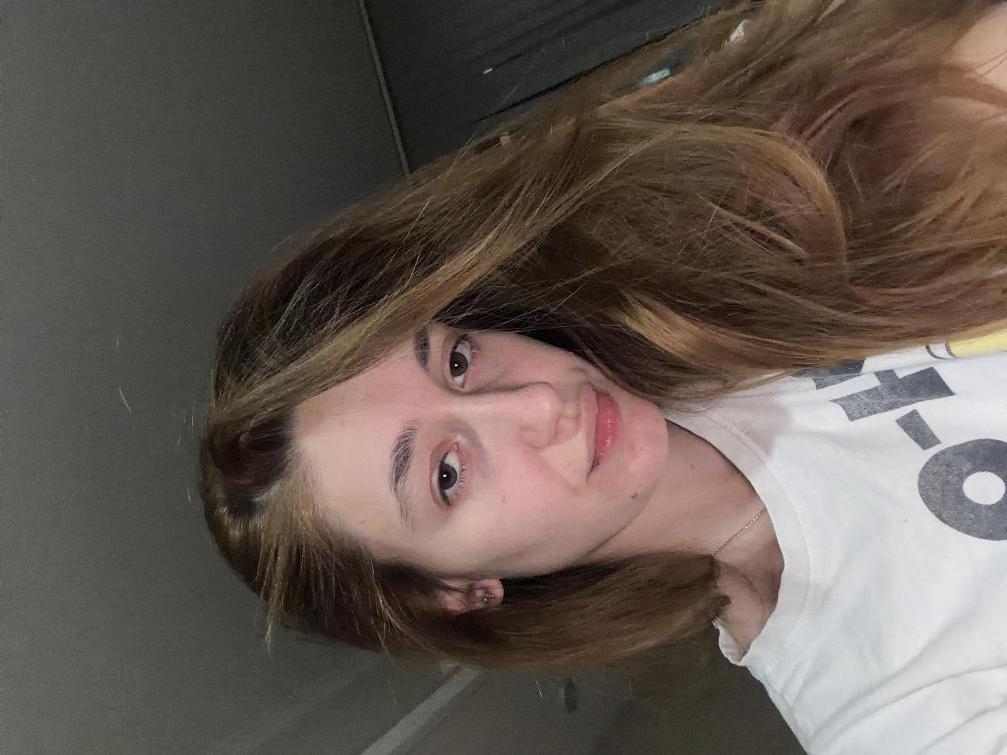 A young woman with long brown hair is the subject of a selfie. Her face is angled toward the camera with a slight smile. She is wearing a white t-shirt with a large, partially obscured gray graphic. A thin, delicate necklace is visible around her neck. The background consists of a gray wall. The location cannot be determined from the image.