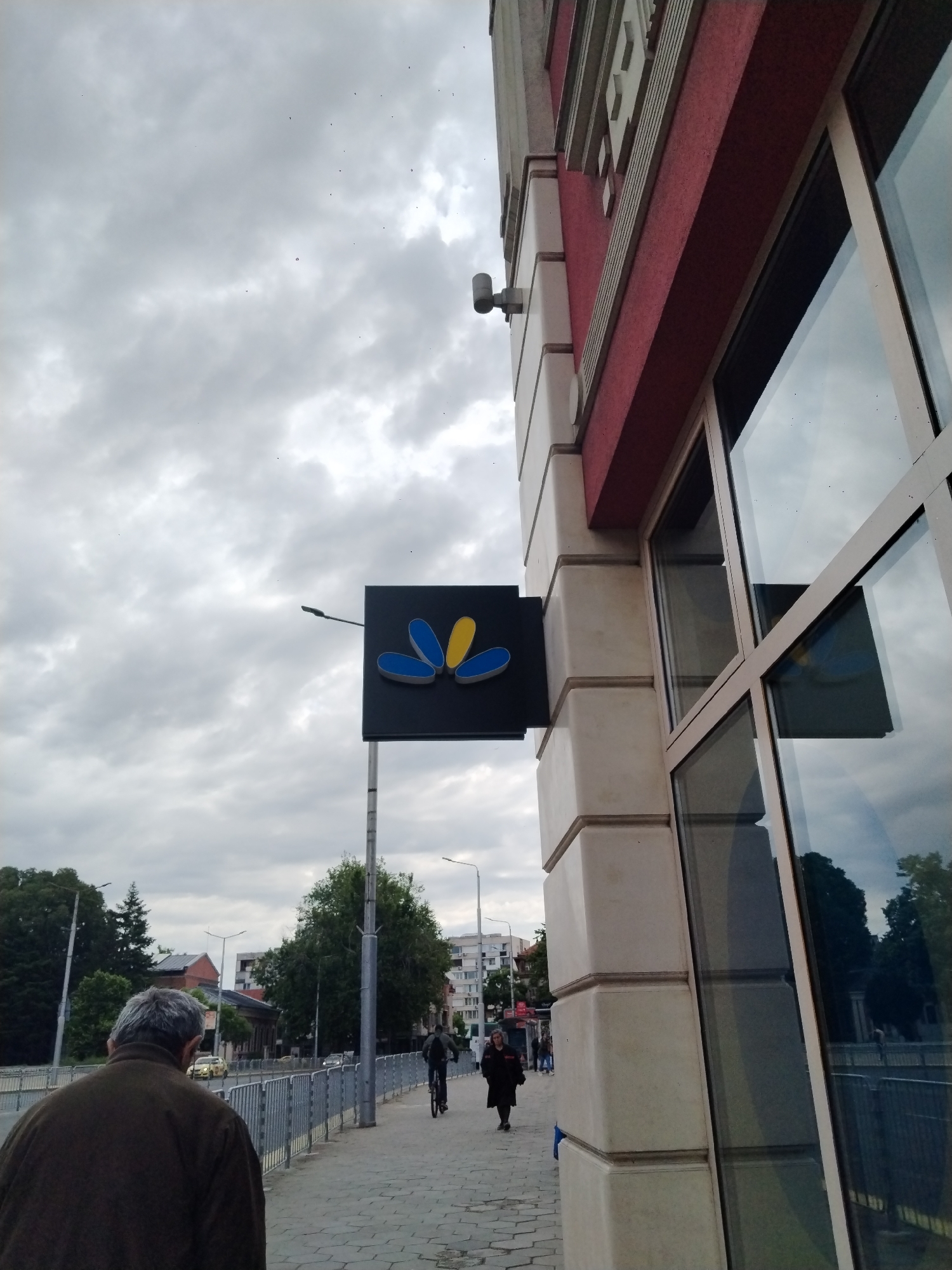 The image depicts a street scene in Plovdiv, Bulgaria, under a cloudy sky. A sign with a blue and yellow flower logo is attached to the corner of a building with cream-colored stone detailing and large windows. The building has a red stripe near the roof line. A pedestrian walkway runs along the building, with a low metal fence separating it from the street. A man with gray hair is walking away from the camera in the foreground, wearing a brown jacket. Further down the walkway, a person is riding a bicycle and another person is walking towards the camera. In the background, trees, buildings, and streetlights are visible.
