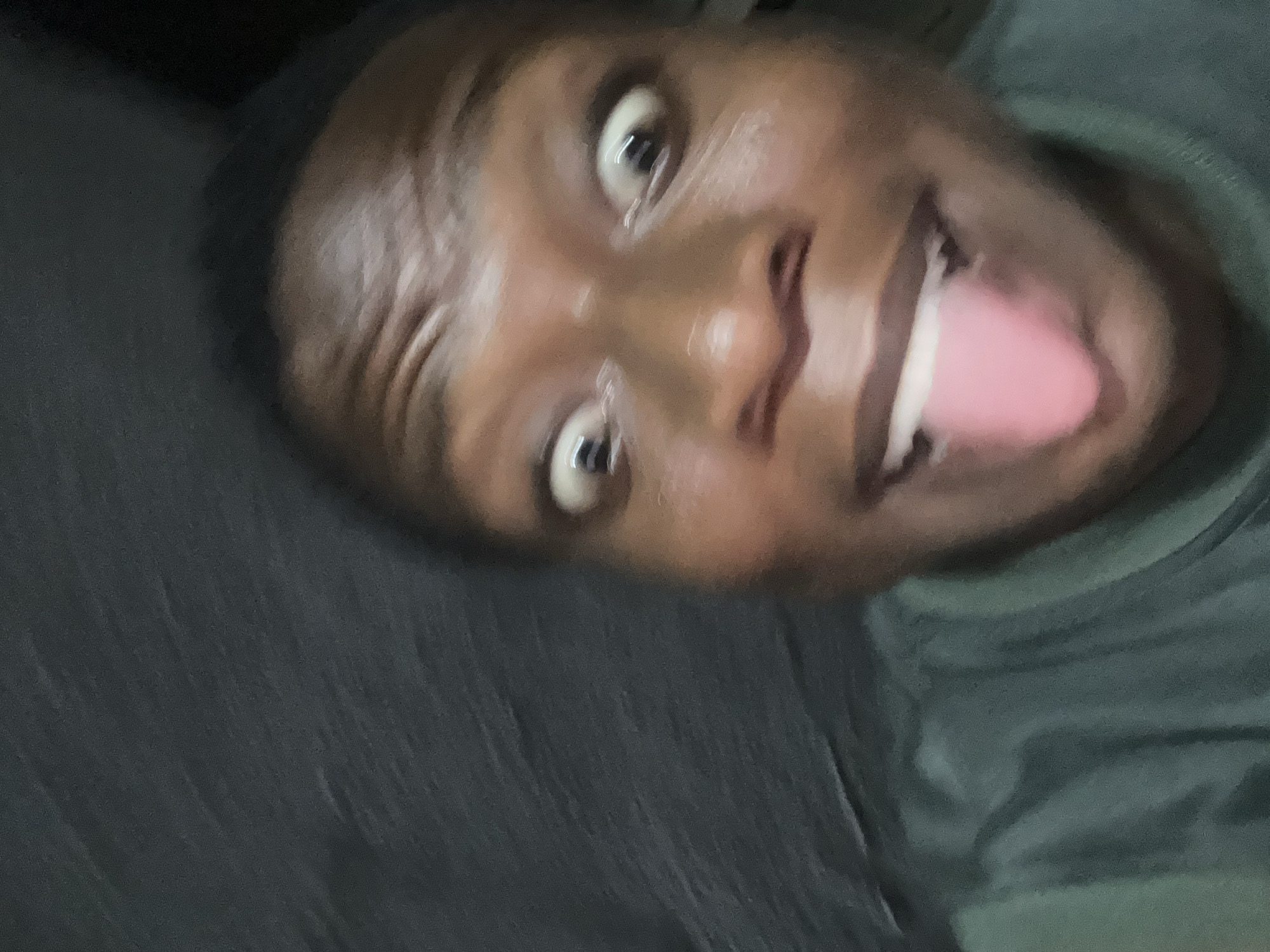 A close-up, slightly blurred portrait of a Black male facing the camera. He has dark hair and skin, with visible moisture on his forehead. His eyes are wide open and expressive. His mouth is open with his tongue sticking out. He is wearing a dark green shirt. The background is blurry and indistinct, appearing to be a dark-colored fabric or surface. The location is unknown.