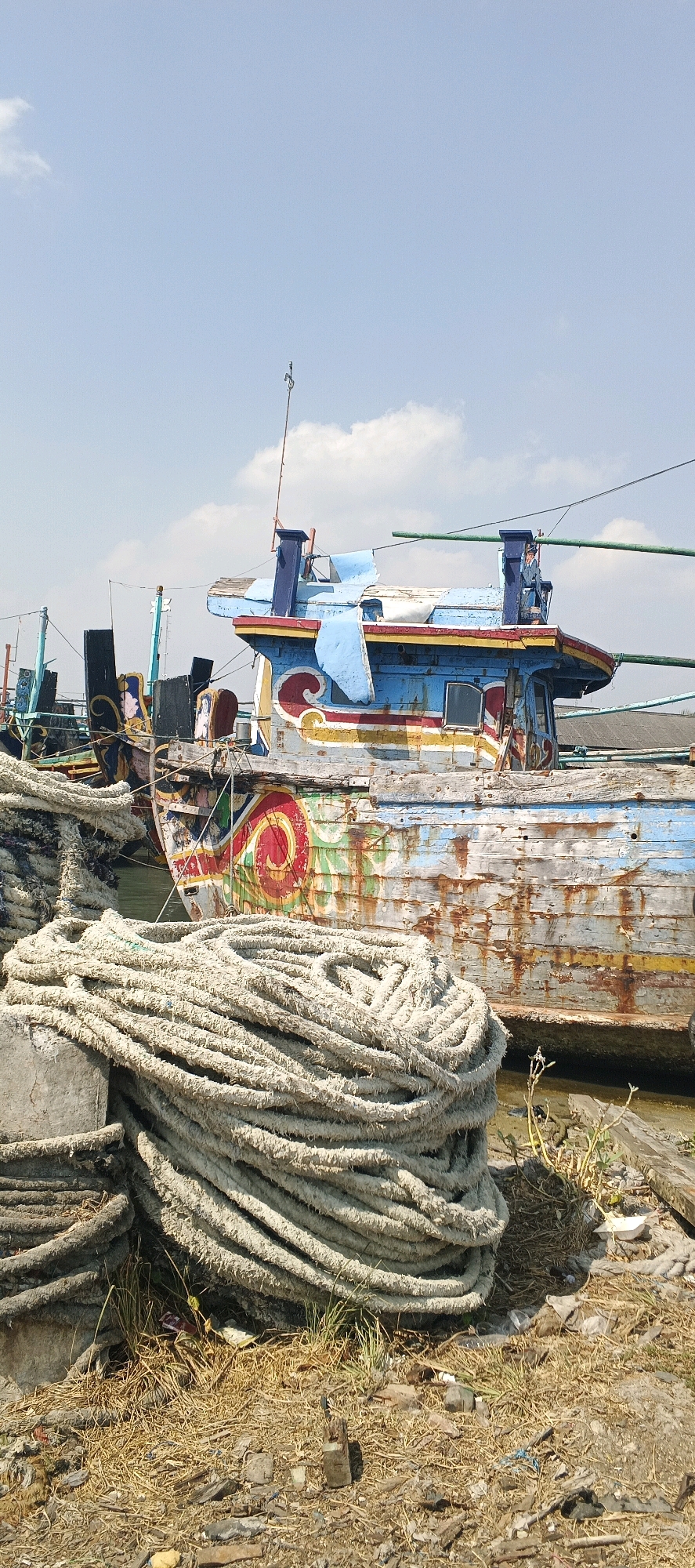 A weathered, brightly painted fishing boat is docked in shallow water, its hull showing signs of rust and peeling paint. The boat features a blue superstructure with red and yellow trim, adorned with decorative patterns. In the foreground, large coils of thick, worn rope are piled on the shore, alongside dry grass, debris, and scattered rocks. The sky above is blue with scattered white clouds. The scene appears to be a port or harbor in Blimbing, Indonesia, characterized by maritime activity and the presence of fishing vessels. There are no people visible in the image.