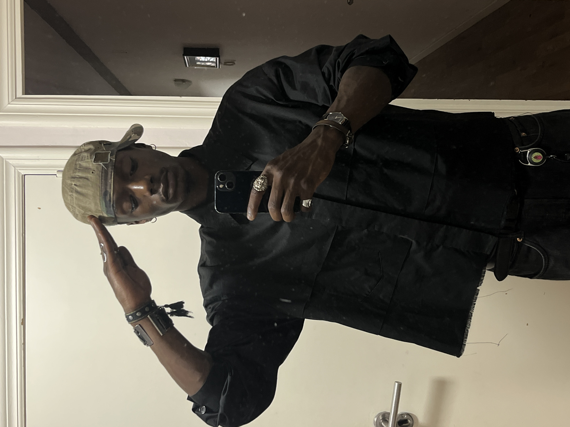 The image is a mirror selfie taken indoors in what appears to be a room in Barcelona, Spain. The primary subject is a person with dark skin, facing forward and looking directly into the camera/mirror. They are wearing a light-colored, possibly khaki or camouflage-patterned baseball cap backward, a black long-sleeved utility or workwear-style shirt with a frayed hem, and black pants or jeans.
The person is holding a black smartphone in their right hand, using it to capture their reflection. They are adorned with several accessories: on their left wrist, a watch with a dark strap and a circular, silver-toned face, along with a studded black leather cuff bracelet and a wider, metallic silver-toned cuff bracelet. On their right hand, a prominent ring with an ornate design (resembling a lion's head or similar motif) is worn on the ring finger, and a plain silver ring is on the pinky finger. Small silver rings are also visible on the fingers of the left hand. Attached to their pants on the right side is a small, round, metallic object, possibly a retractable keyring or badge holder.
The background visible in the mirror reflection shows a light-colored wall with white molding, and a dark ceiling with a rectangular light fixture. A silver plumbing fixture (possibly a tap or toilet roll holder) is visible in the lower part of the reflection. The person's expression is neutral, and the indoor lighting appears somewhat dim. There is no visible text in the image.