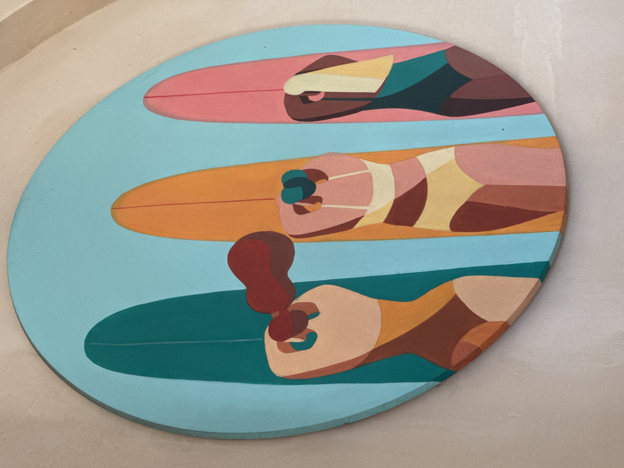 This is a photograph of a circular painted image depicting three people lying on surfboards in the ocean. The backdrop is light blue, representing the ocean. The top figure is on a pink surfboard, the middle figure is on an orange surfboard, and the bottom figure is on a green surfboard. The people are depicted in a stylized, geometric manner. It's located in Kuta Selatan, Indonesia.