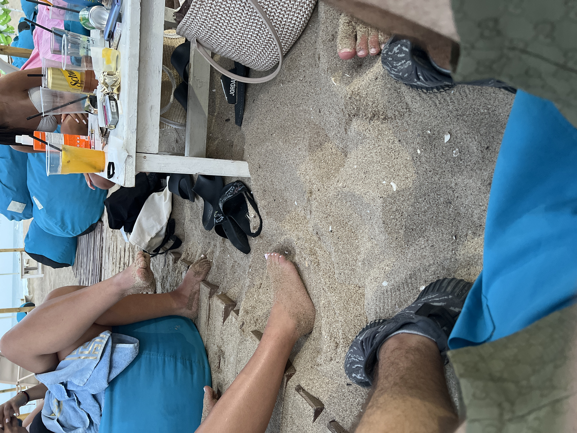 The image depicts a sandy beach environment in Obzor, Bulgaria, likely during daylight hours. The scene is focused on a group of people relaxing on beanbag chairs and around a low table. Several individuals' legs and feet are visible, some adorned with sandals or flip-flops. One person is partially covered by a light blue towel. On the white wooden table, there are multiple plastic cups with straws, some containing beverages, and a can of Schweppes. A pack of cigarettes and a mobile phone are also present. Scattered on the sand around the table are additional pairs of flip-flops and a woven handbag. The background suggests an outdoor seating area with more beanbag chairs and glimpses of beach umbrellas and the sea.