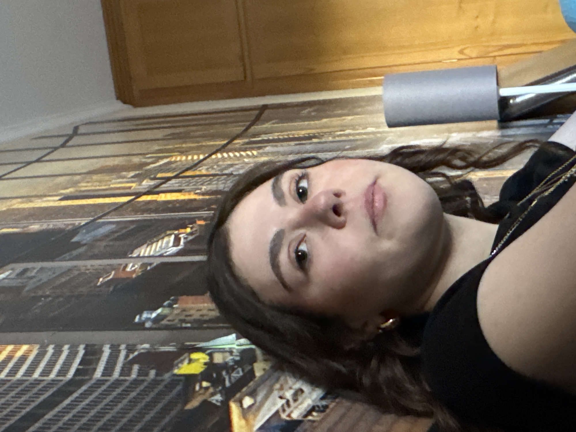 A person is lying down on a surface decorated with an image of a city, giving the impression they are on the floor. The individual has brown hair and is wearing a black top. A cylindrical object, potentially a foam roller or similar item, rests near their head. The background includes what appears to be wooden cabinetry or paneling. The image does not include any identifiable details related to Mirandela, Portugal.