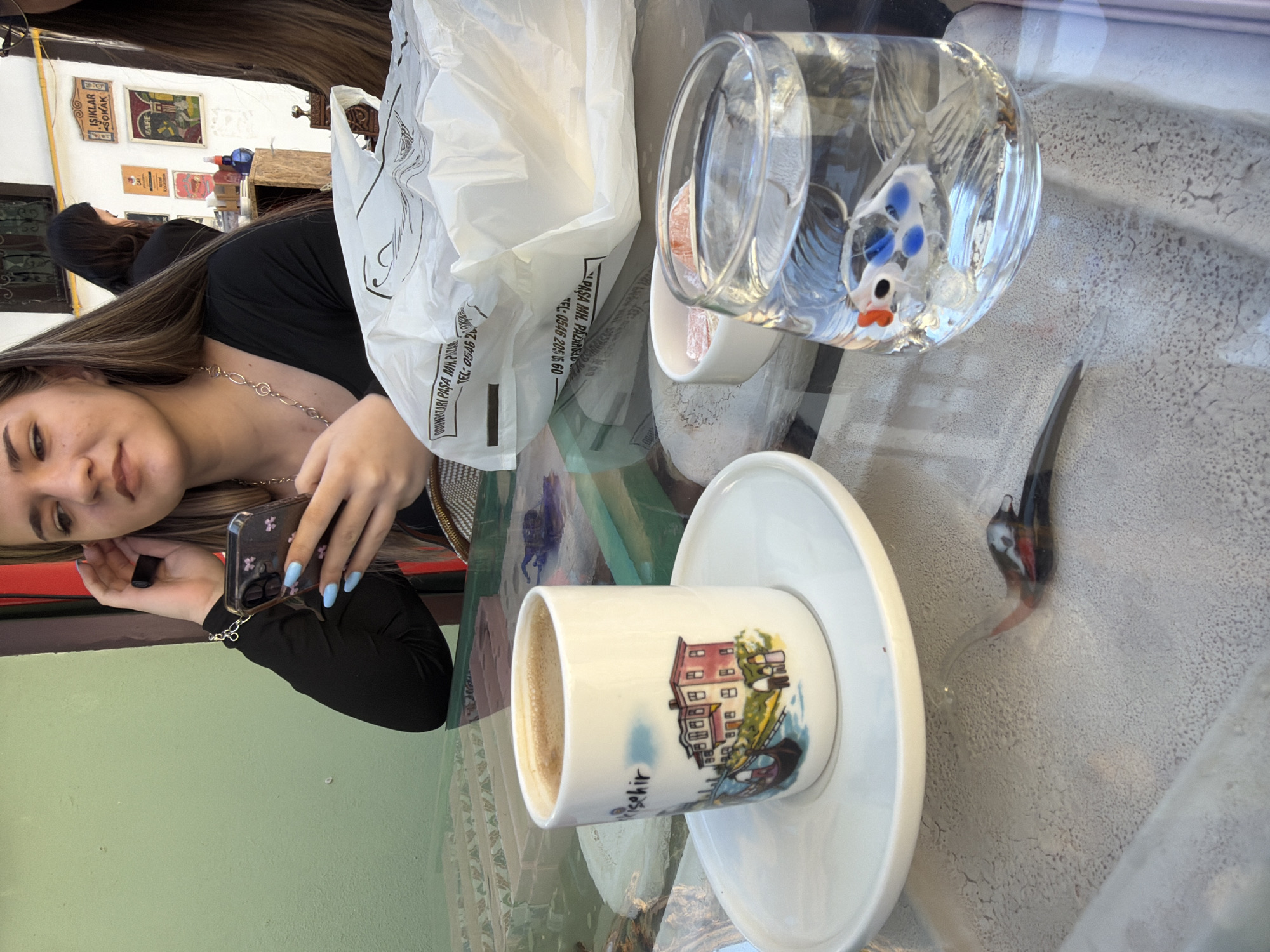 The image captures a casual cafe setting, likely outdoors or semi-outdoors, focusing on a young woman and items on a glass table in Eskisehir, Turkey.
The primary subject is a young woman with long, straight brown hair, wearing a black long-sleeved top and a silver chain necklace. She looks directly at the camera with a subtle smile or neutral expression, holding a smartphone with a floral pattern case in her hands. Her fingernails are painted a light blue color. Another person with dark hair is partially visible in the background, seated and facing away from the camera.
On the glass table, there's a small white cup of Turkish coffee with a colorful illustration of buildings and the word "Eskisehir" (partially visible as "sehir") on its side, resting on a white saucer. Next to it, a clear glass filled with water contains a decorative glass fish with blue spots and red fins. A small white bowl beside the water glass holds several pieces of pinkish Turkish delight. A white plastic shopping bag with black text is also on the table. A small, dark-colored bird figurine or ornament with red and white accents is visible on the table's surface.
The background features a light green wall in the foreground, transitioning to a white wall adorned with several framed pictures or posters. Text visible on these posters includes "IŞIKLAR SOKAK" (Işıklar Street), "ÇAY" (Tea), and "KAHVE" (Coffee), suggesting a cafe. The plastic bag displays text like "DOĞANLAR PAŞA MH. PAZAR" (Paşa Neighborhood Market) and a telephone number, reinforcing the Turkish location.
The scene is brightly lit, indicating daytime, possibly sunny, creating a relaxed atmosphere. The woman appears to be enjoying a coffee break, perhaps in a historic or quaint part of Eskisehir.
