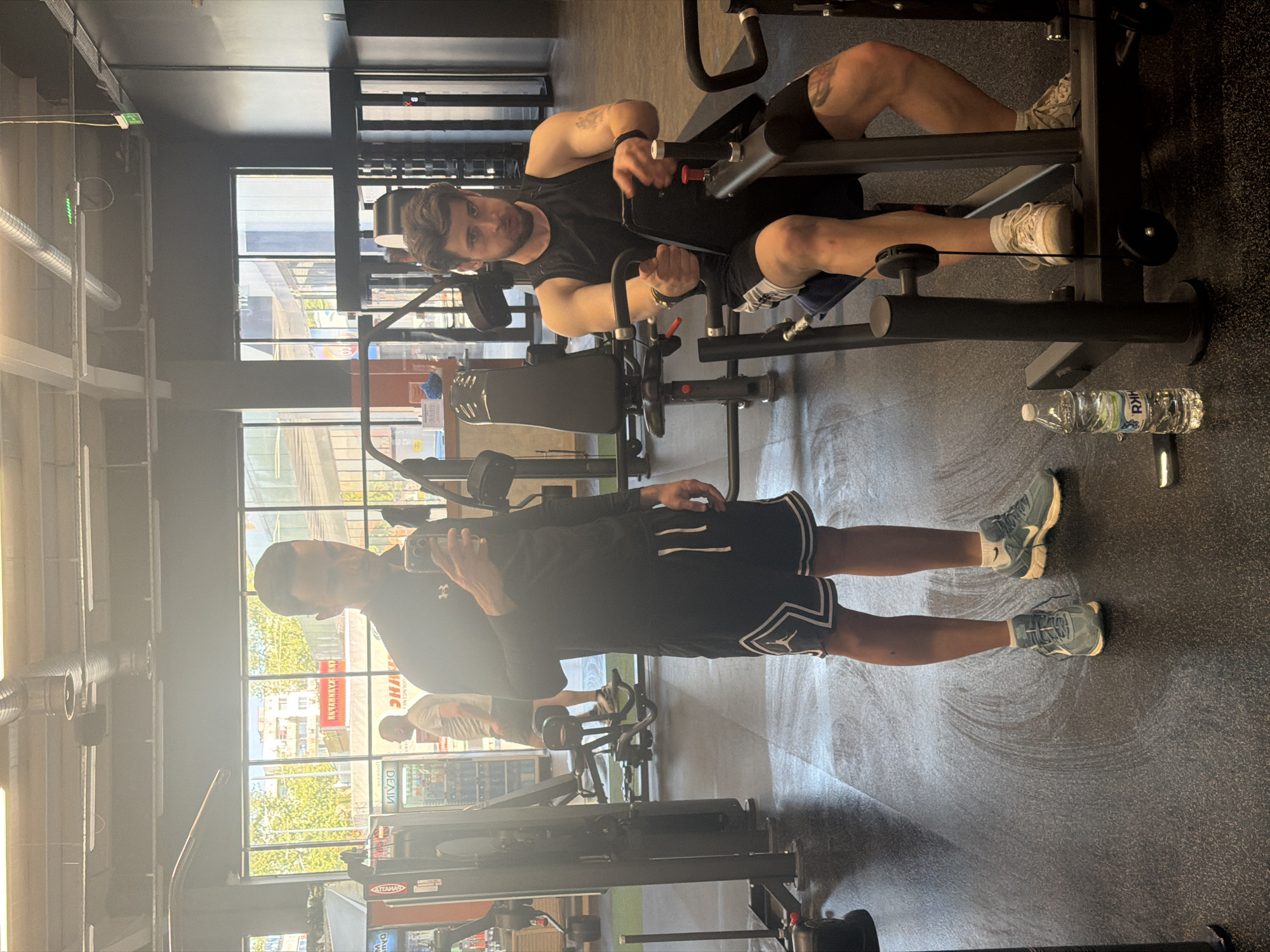 The image depicts a gym interior. A man is exercising on a seated cable row machine; he has a beard, dark hair, and a tattoo on his left arm, wearing a black tank top and shorts. Another man is standing in front of him, holding a phone, potentially taking a picture. He is wearing a black top and shorts with white trim. A water bottle lies on the floor next to the machine. Outside the window are buildings. A building across the street has a sign in Cyrillic, reading “ХРАНИТЕЛЕН МАГАЗИН”. The address of this shop is 33 Hristo Botev Blvd., Plovdiv, Bulgaria.