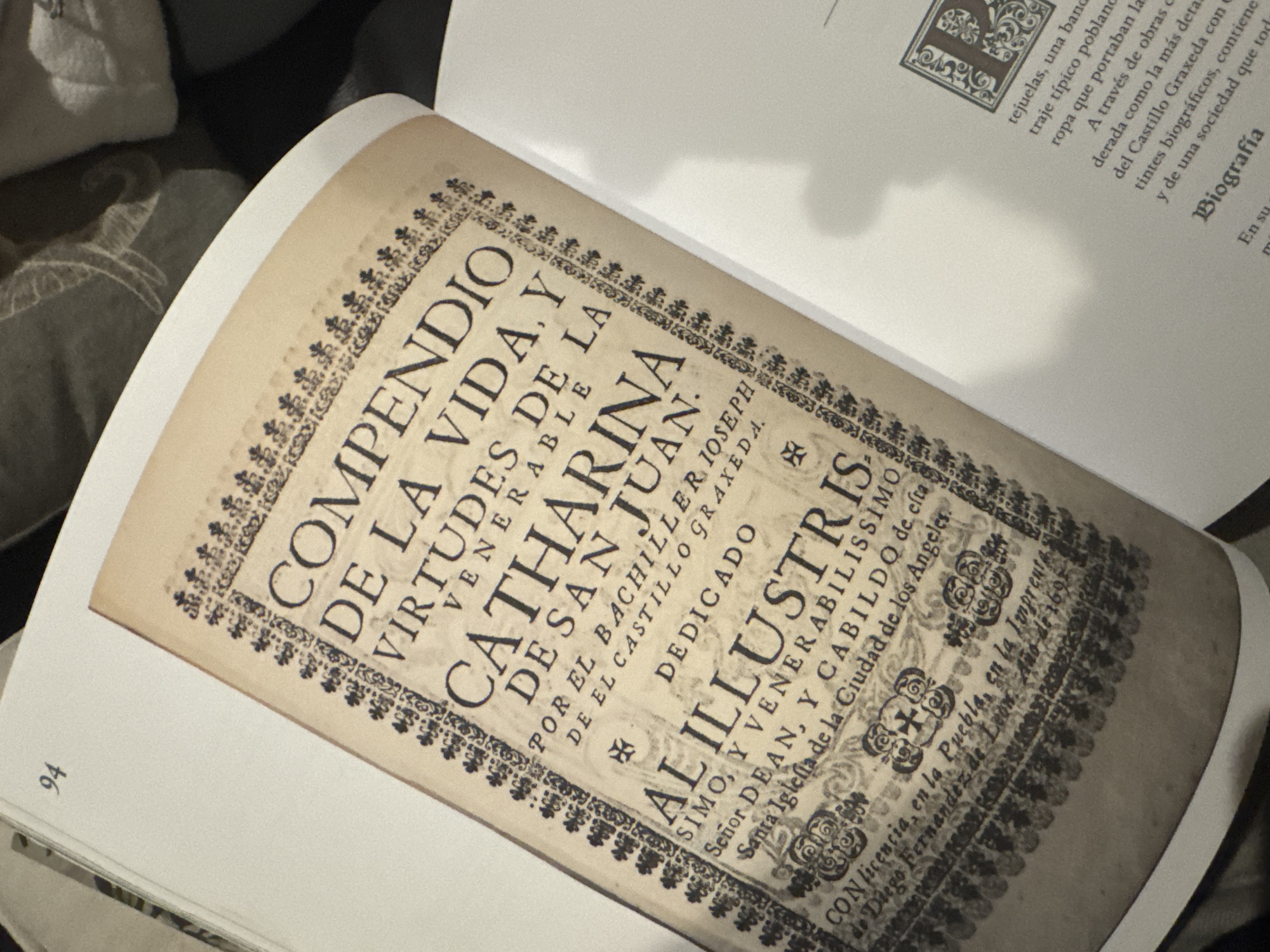 A close-up photograph reveals a page in a book, showcasing a title page. The title reads "COMPENDIO DE LA VIDA. Y VIRTUDES DE LA VENERABLE CATHARINA DE SAN JUAN," followed by "POR EL BACHILLER IOSEPH DE EL CASTILLO GRAXEDA. DEDICADO." Beneath this, the text continues "AL ILLVSTRIS SIMO, Y VENERABILISSIMO Señor DEAN, Y CABILDO de esta Santa Iglesia de la Ciudad de los Angeles,". The page specifies that the printing took place "CON licencia en la Puebla, en la Imprenta de Diego Fernandez de Leon. Año de 1691." The image is set within the context of a larger book, with text visible on the adjacent page discussing Puebla. A numerical "94" is seen at the bottom left, likely indicating a page number. The lighting suggests an indoor environment.