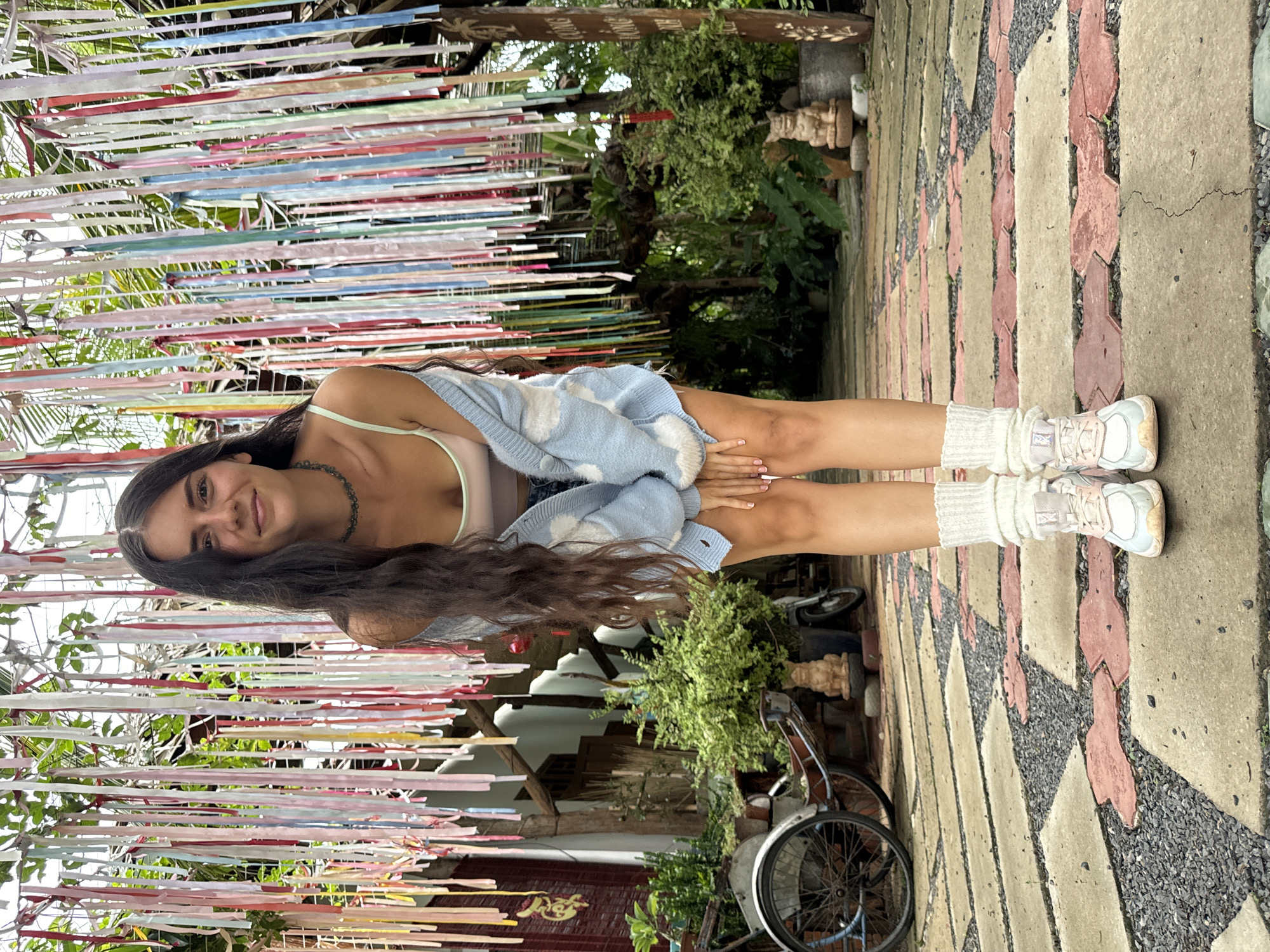 A young woman stands on a tiled walkway in what appears to be an outdoor courtyard. She is facing the camera with a slight smile. She is wearing a light-colored crop top, a light blue cardigan with cloud patterns, white leg warmers, and light-colored athletic shoes. Her hair is long and dark.
Overhead, there is a canopy of multi-colored ribbons. Behind her, there is lush greenery, suggesting a garden or tropical environment. To her left, there is a bicycle parked near a building with decorative elements. The location is Mũi Né, Vietnam.