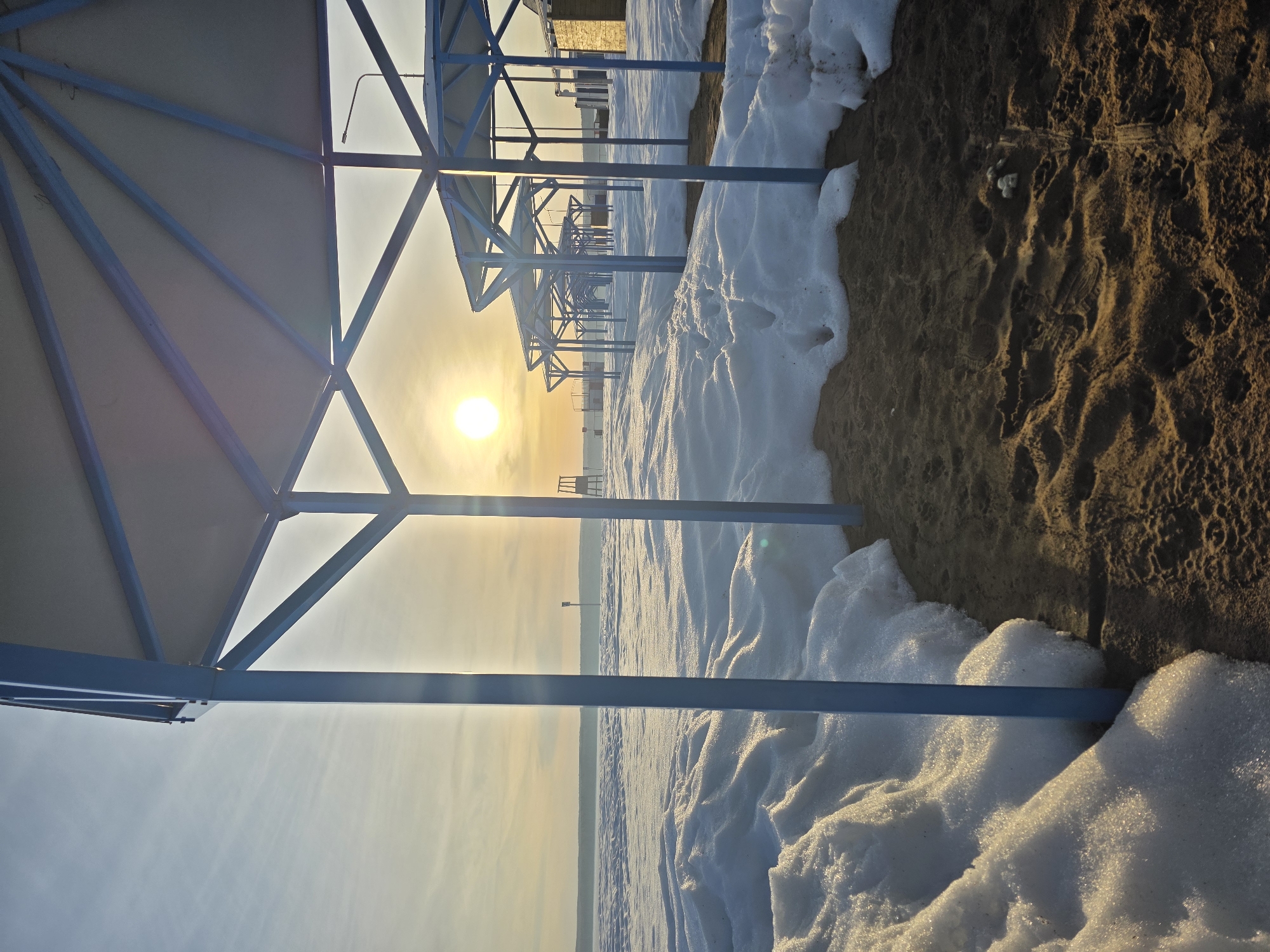 The image depicts a snow-covered beach scene in Chaikovsky, Russia, likely during winter. A row of beach umbrellas with blue frames is visible, receding into the distance. The sun is low on the horizon, creating a strong glare. Patches of snow cover the sand on the right side of the image. The sky appears hazy.