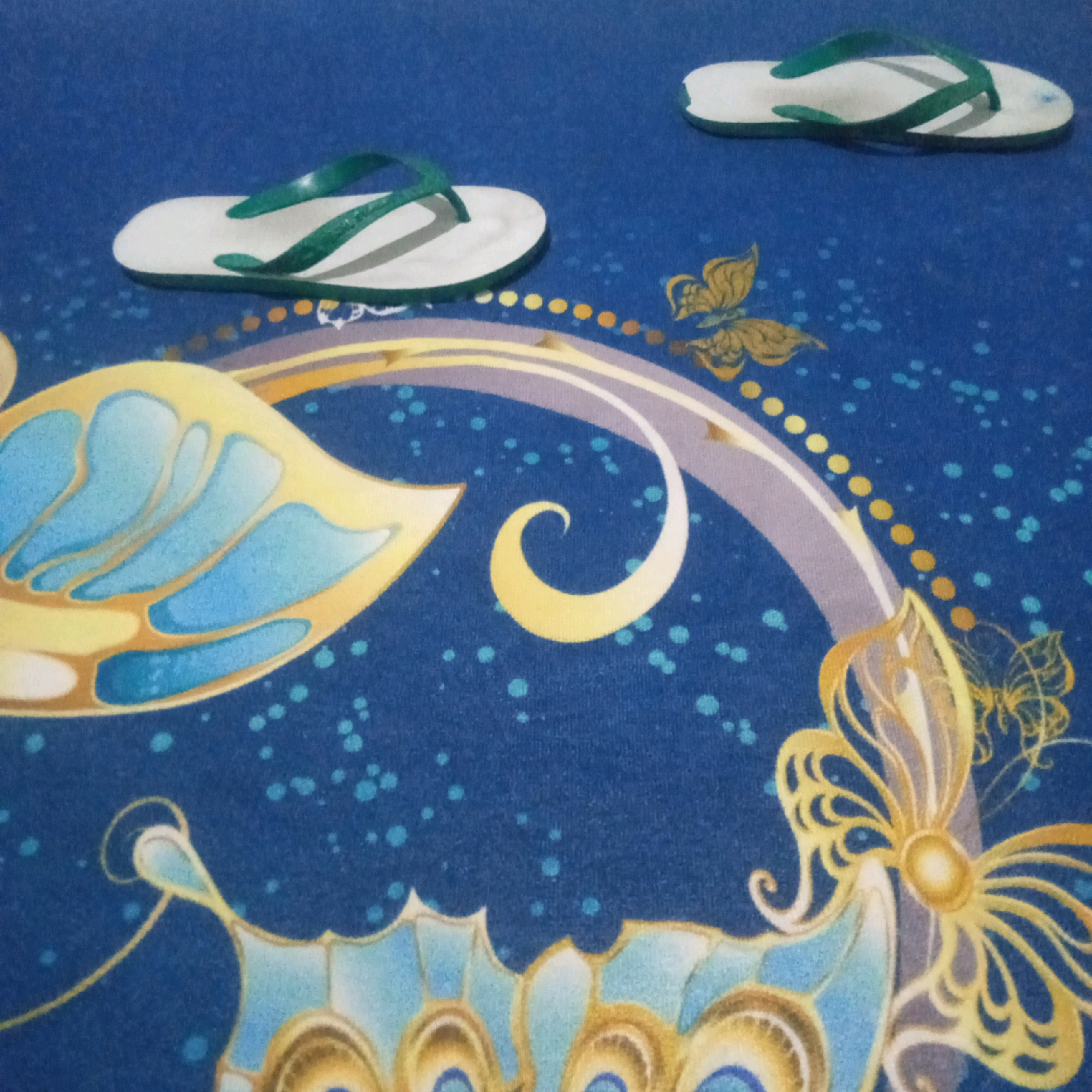 Two white flip-flops with green straps are placed on a dark blue surface. The surface is decorated with a colorful, abstract design featuring yellow and blue butterfly-like shapes, swirling lines, and small blue and yellow dots. The flip-flops are positioned in the upper portion of the image, with one on the left and another on the right. The overall scene appears to be indoors or on a protected surface, likely in a residential setting in Kendayakan, Indonesia, given the context.