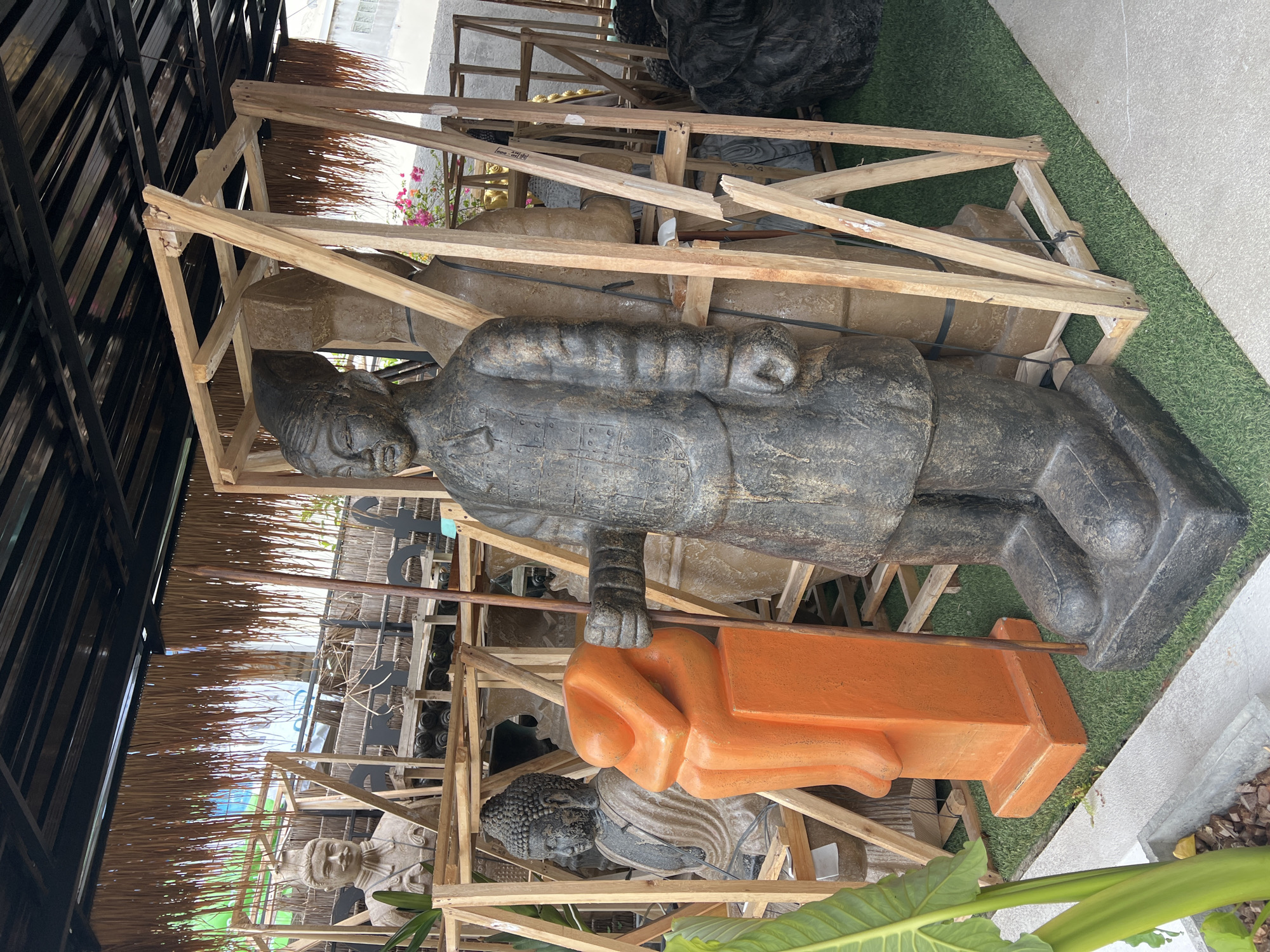The image shows a collection of statues in Baan Bo Phut, Thailand, contained within a wooden framework. Prominently displayed is a gray-colored statue of a man in traditional garb, lying down with its right hand resting on an orange platform. Below this statue are smaller statues of Buddha, also within the wooden framework. The scene is partly enclosed by a thatched roof structure. The environment is composed of faux grass and a concrete pathway, suggesting an outdoor or partially covered location.