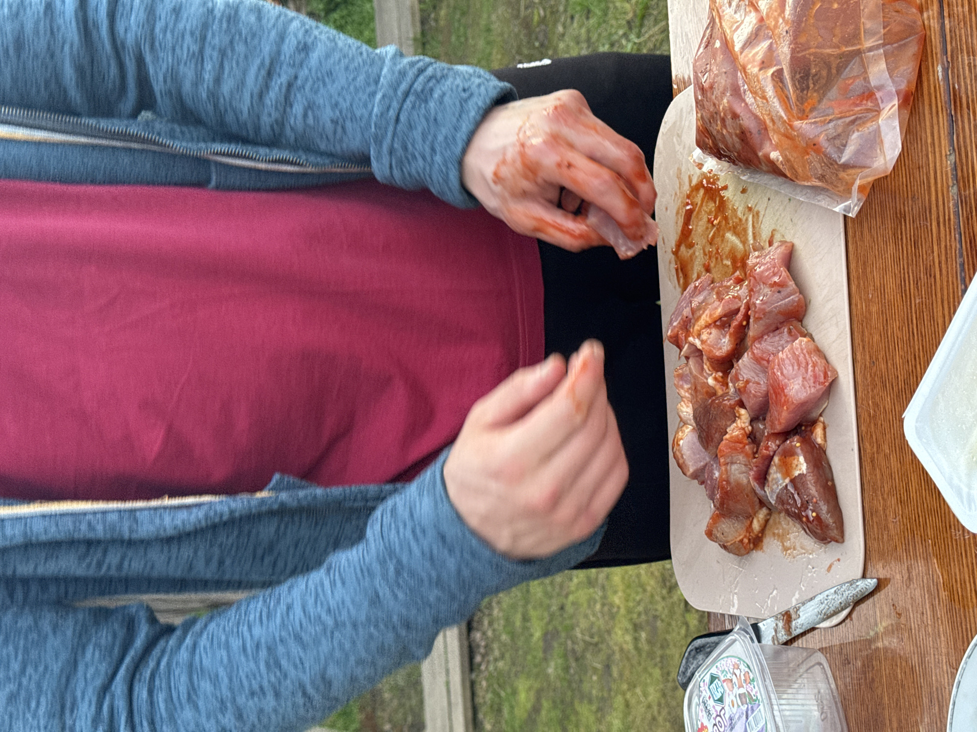 An outdoor scene, possibly in Maksimovo, Russia, features a person wearing a blue hooded jacket over a pink t-shirt. Their hands, stained with a reddish sauce, are actively engaged with raw meat. A cutting board displays several pieces of marinated meat, while a plastic bag on the right also contains marinated meat. A small plastic container and a knife are also present on a wooden table, set against a backdrop of greenery.