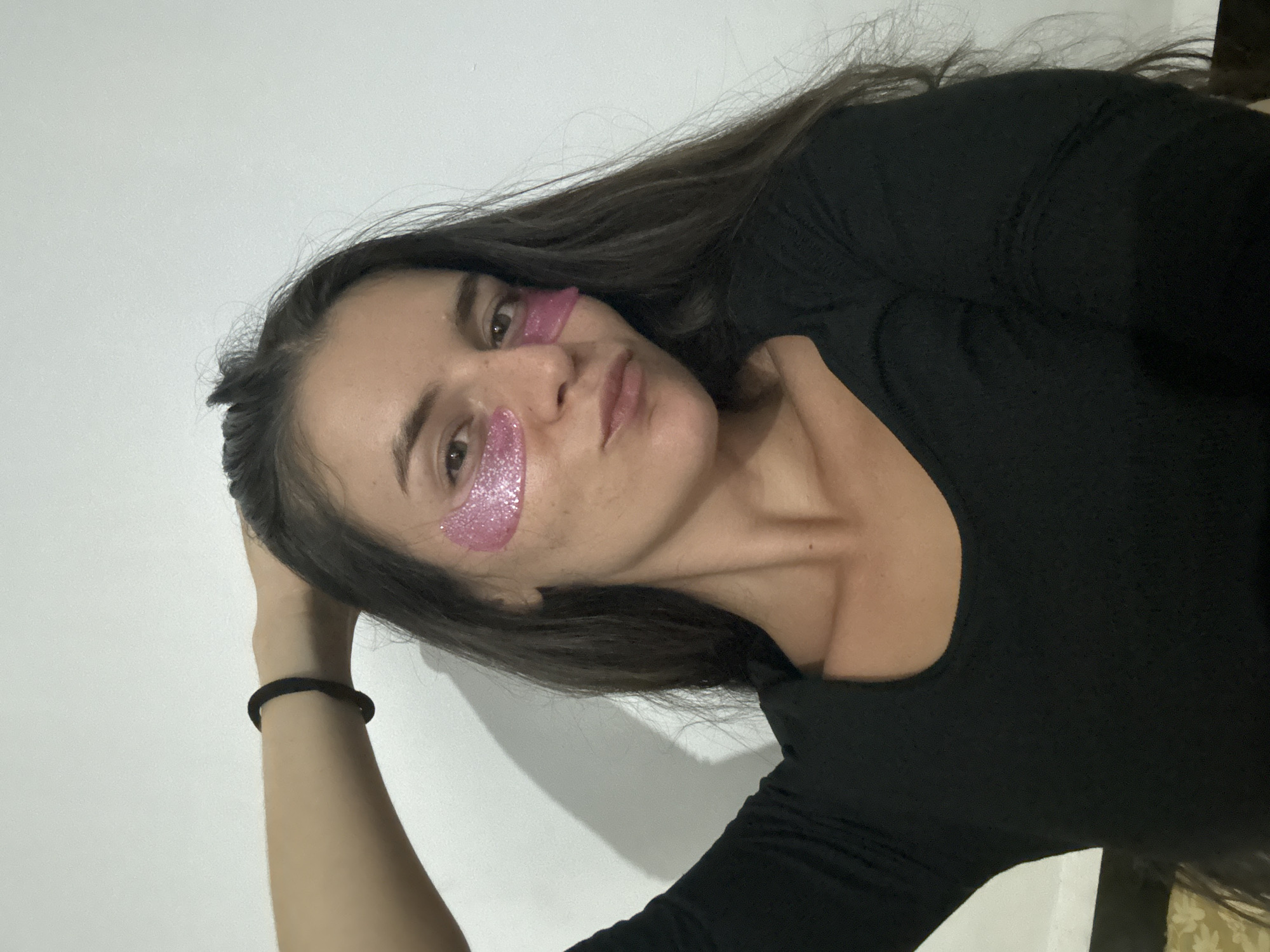 A young woman, likely in her late teens or early twenties, is captured in a selfie taken indoors against a plain white wall. She has long, dark brown hair that cascades around her shoulders and frames her face. She is wearing a black, long-sleeved top. Her face is adorned with two shimmering pink under-eye patches, adding a playful and cosmetic touch. Her expression is relaxed, with a slight pout. Her left arm is bent, with her hand resting on the back of her head, and a black hair tie is visible on her wrist. The lighting suggests it is daytime, and the overall scene appears casual and personal. There is no visible text in the image. The context suggests this photo was taken in Mũi Né, Vietnam.