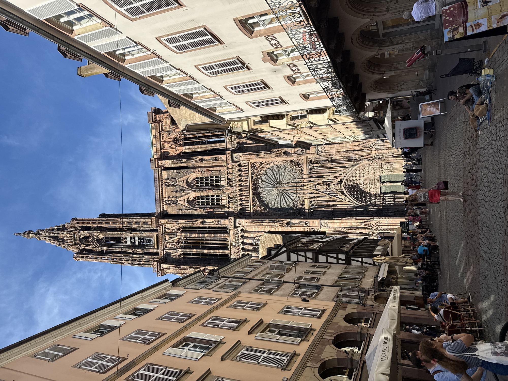 The image depicts a view of the Strasbourg Cathedral, officially known as the Cathédrale Notre-Dame de Strasbourg, on a sunny day. The cathedral's imposing Gothic facade, constructed from pink sandstone, dominates the background with its intricate carvings, a large rose window, and a tall spire. The foreground and sides are lined with narrow, multi-story buildings in pastel colors, typical of Strasbourg's architecture, featuring shuttered windows and pitched roofs. A cobblestone street runs through the center, populated by pedestrians. Several people are seated at outdoor tables of a cafe under umbrellas, enjoying meals and drinks. Others are walking along the street, some in casual attire, and a few are gathered near street vendors. A man is sitting on the ground with two dogs. The overall scene is a vibrant streetscape in the historic center of Strasbourg, France, capturing the grandeur of the cathedral juxtaposed with everyday life.