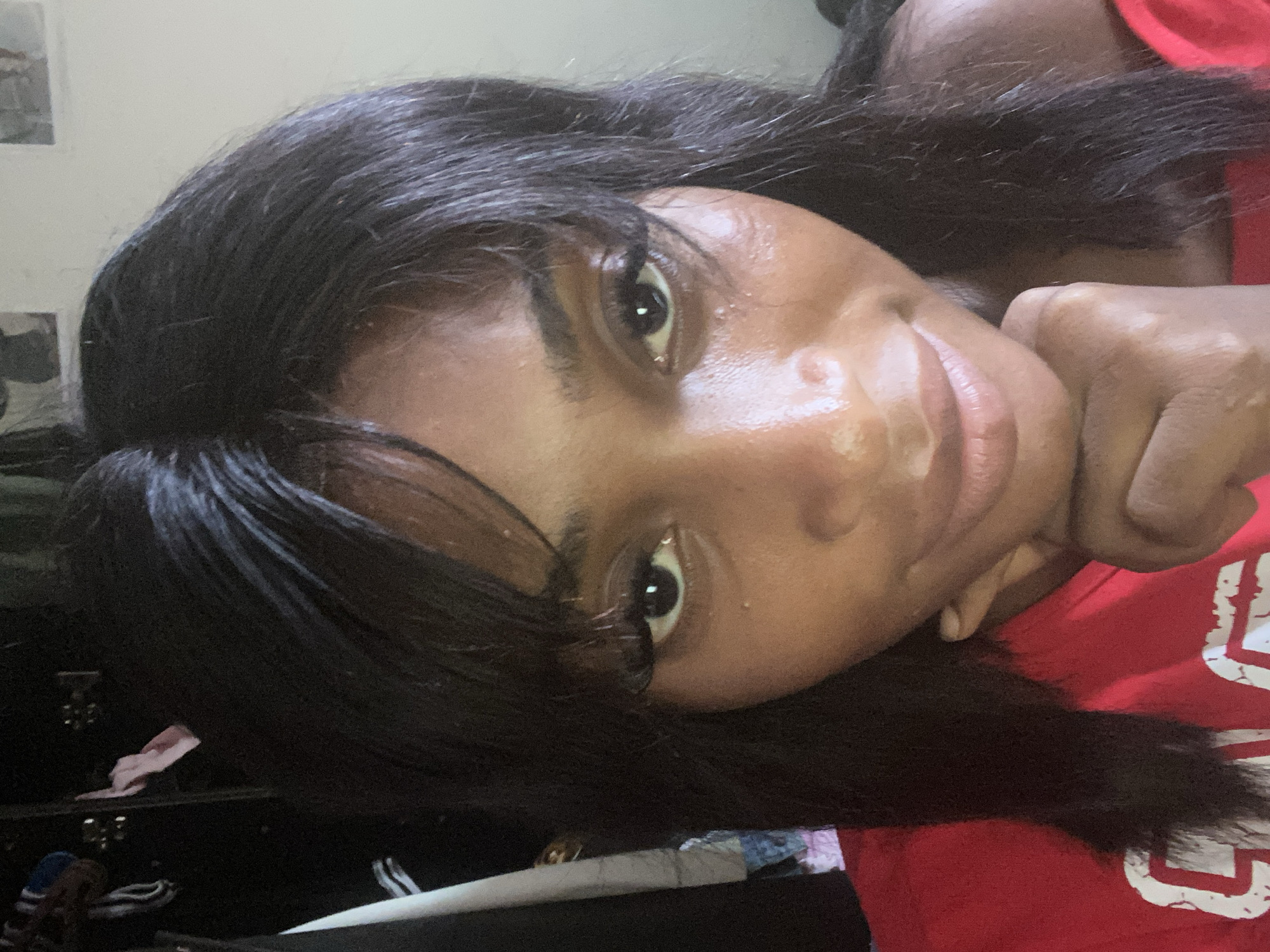 A close-up, head-and-shoulders selfie shows a Black woman with long, dark hair and bangs. She is wearing a red shirt and resting her chin on her hand. Her eyes are open and she is looking towards the camera. Her skin appears to be slightly shiny, possibly from sweat or makeup. The background is dark and appears to be indoors, possibly a room with clothing visible in the upper left corner. There are no discernible features that would indicate a specific location in Kubwa, Nigeria.