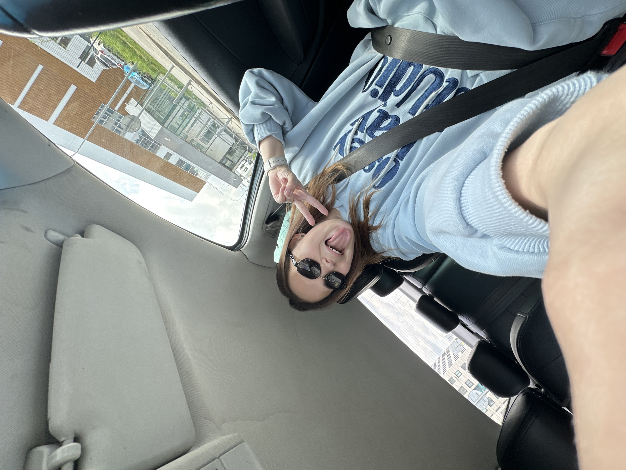 A person is pictured in a car, wearing a light blue sweatshirt and sunglasses. They are also wearing a seatbelt and making a peace sign with their fingers while sticking out their tongue. The buildings outside the car appear to be of modern construction. Based on the exterior architecture, this image was taken in Perm, Russia.