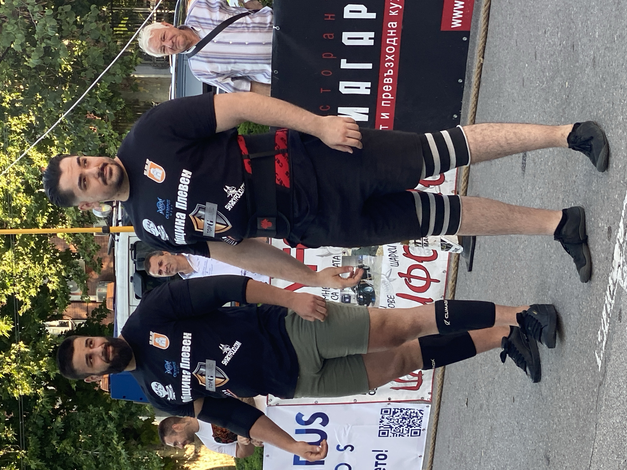 Two muscular men in athletic attire stand on an asphalt surface, possibly outdoors. Both are wearing black t-shirts with logos and text in Bulgarian, indicating a connection to "Община Плевен" (Municipality of Pleven). The man on the left wears khaki shorts and black leg sleeves. The man on the right wears black shorts, a black weightlifting belt with red accents, and striped leg sleeves. In the background, there are trees, a white vehicle, and banners with Bulgarian text. The overall setting suggests an event or competition taking place in Pleven, Bulgaria.