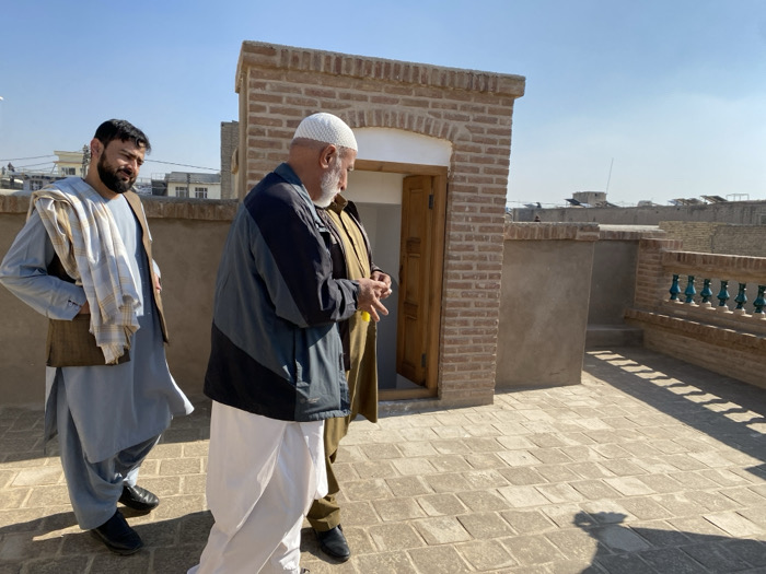 The image displays three adult males on a tiled rooftop in Kandahar, Afghanistan, under a clear blue sky. The man on the left, with a dark beard, wears a light blue shalwar kameez, a brown vest, and a light-colored plaid scarf draped over his left shoulder; he looks towards the right with his hands in his pockets. The central figure, an older man with a white beard, wears a white skullcap, a dark blue and black patterned jacket, and white shalwar pants; he appears to be looking downwards or at his hands. A third man, largely obscured behind the central figure, is partially visible wearing a brown or olive-green traditional outfit.
The rooftop is paved with rectangular, light-colored tiles, exhibiting strong shadows from the bright sunlight. To the right of the men, a brick structure with an arched doorway containing a partially open wooden door is visible. Further to the right, a brick parapet features a decorative green railing with vertical spindles. In the background, a horizon of other urban rooftops and buildings extends under the clear sky.