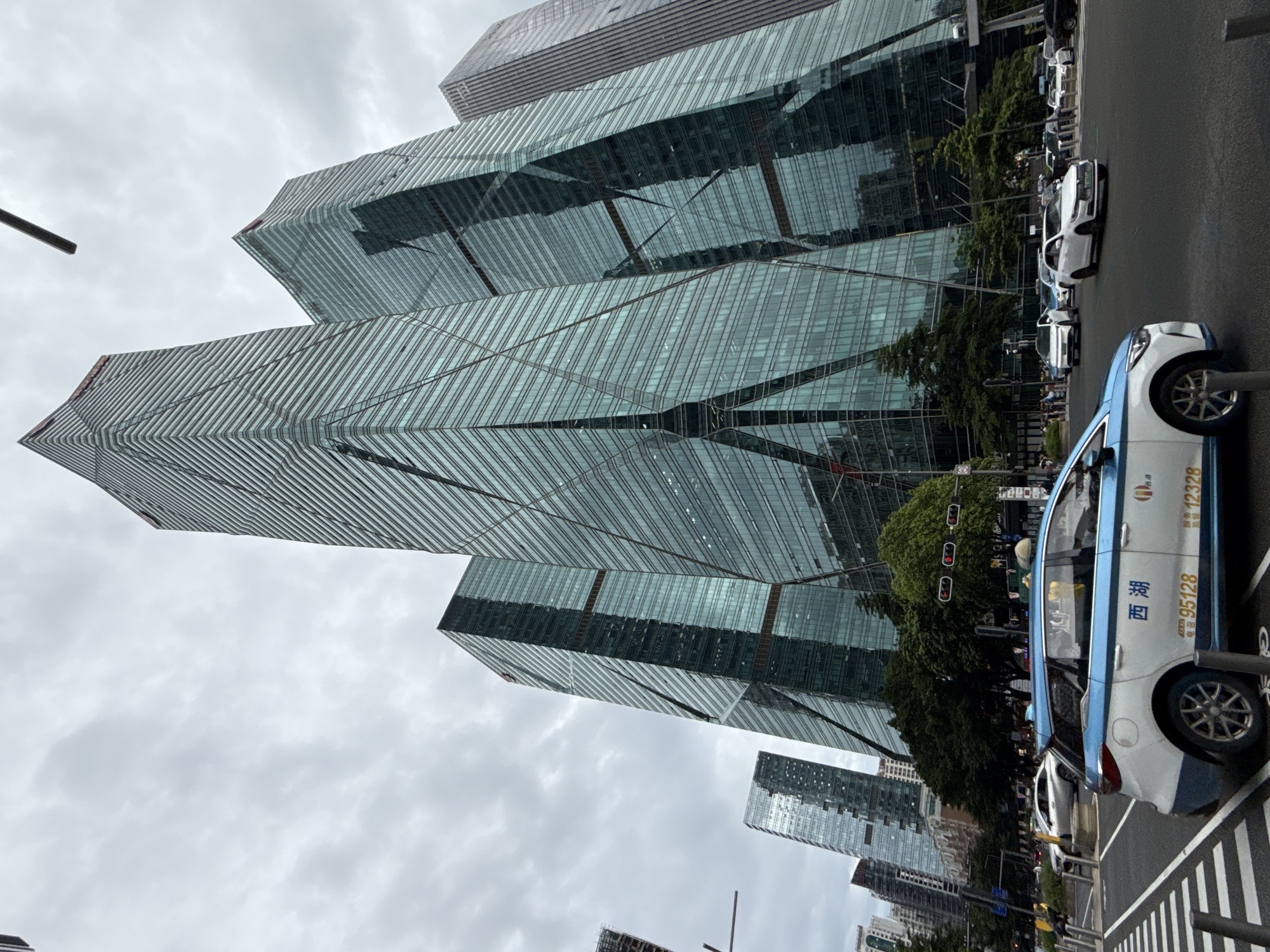 The image shows a street scene in Futian District, China, with several modern skyscrapers dominating the background. The tallest skyscraper has a distinctive angular design with a glass facade. Other buildings in the background are also tall and made of glass, reflecting the cloudy sky. In the foreground, a blue and white taxi with Chinese characters and numbers "95128" and "12328" is parked on the right side of the road. Several other cars are visible on the street, including white sedans and a silver sedan. Pedestrians are also present on the sidewalk. Traffic lights are visible, and the overall atmosphere suggests a busy urban environment.