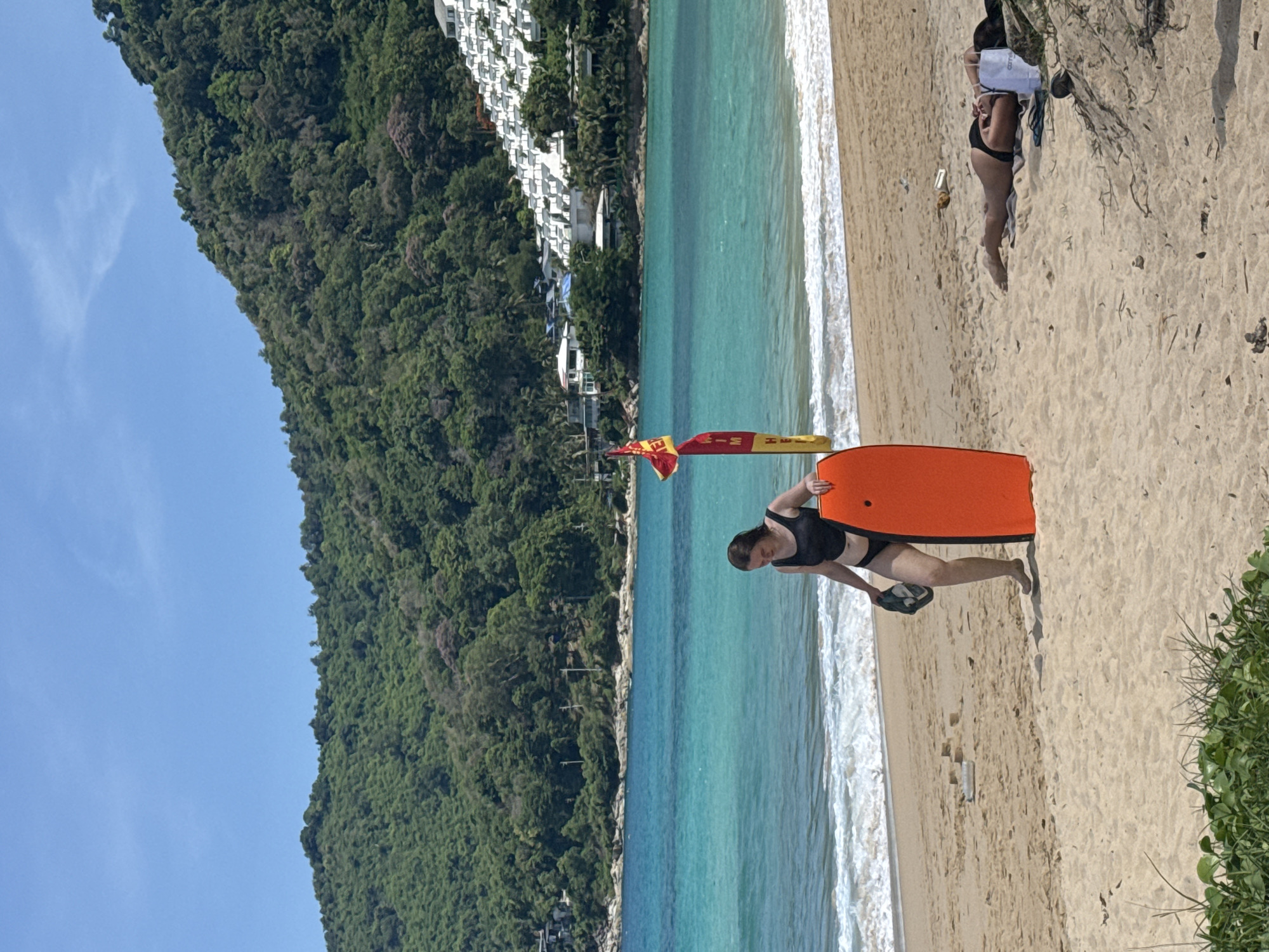 In a medium shot of a sandy beach, a young woman wearing a black swimsuit stands on the left side of the frame holding an orange boogie board and a dark cap. She is facing right, looking down at the boogie board. To her right, a woman in a black bikini lies on her stomach on a towel, facing away from the camera. Behind them, the turquoise ocean stretches to the horizon, with white waves breaking on the shore. In the background, a lush green, tree-covered mountain rises behind the beach. A red and yellow flag with black lettering stands near the water's edge. Several buildings with white triangular roofs are visible on the mountainside. The sky is a clear blue with a few wispy clouds.