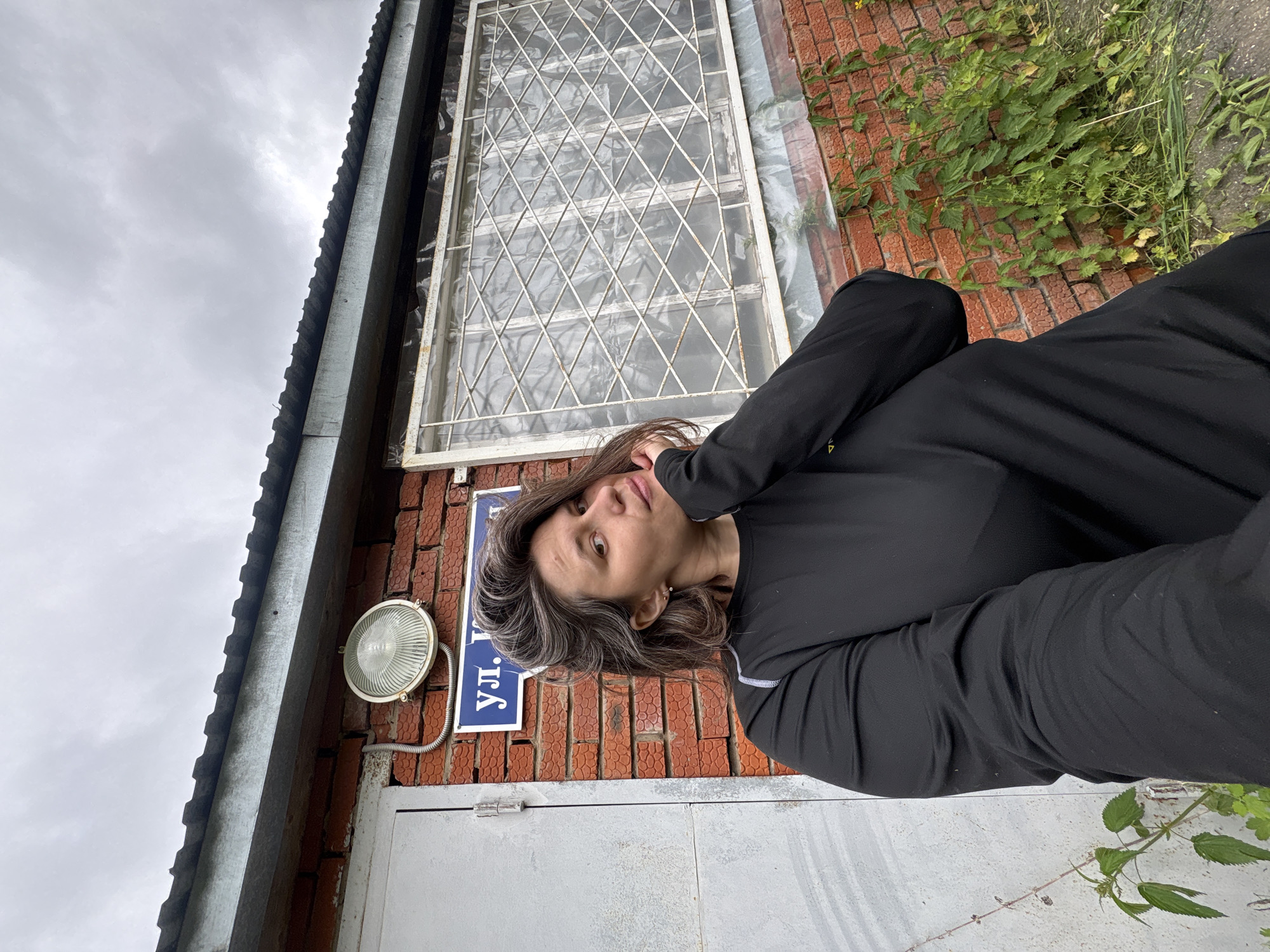 A woman with dark hair and gray streaks poses for a selfie outdoors. She is wearing a long-sleeved black top and is leaning her head on her hand. Behind her is a brick building with a metal roof, a gray door, a round light fixture, and a window with a diamond-patterned metal grate. A blue street sign with white lettering is partially visible. Green plants grow around the base of the building. The sky is overcast and gray.
