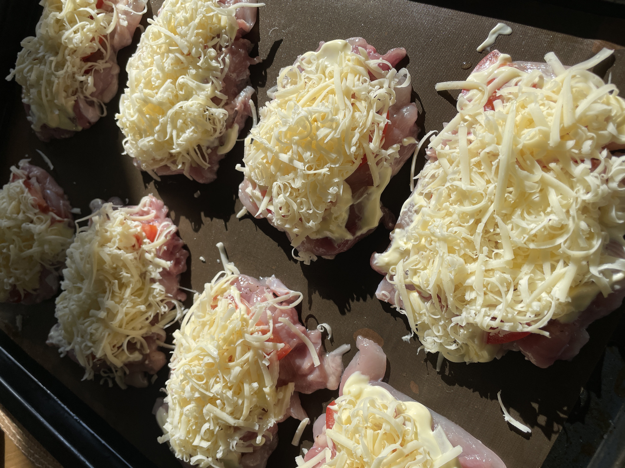 The image shows a black baking tray containing eight pieces of uncooked meat, possibly chicken, topped with shredded cheese and a creamy sauce, likely mayonnaise, and slices of tomato. The meat is arranged in rows on the tray. The scene is brightly lit, suggesting it is taken in natural light. The setting appears to be indoors, with a wooden surface visible in the lower left corner of the image. There are no people in the image. The image does not contain any information linking it to the city of Chaikovsky, Russia.