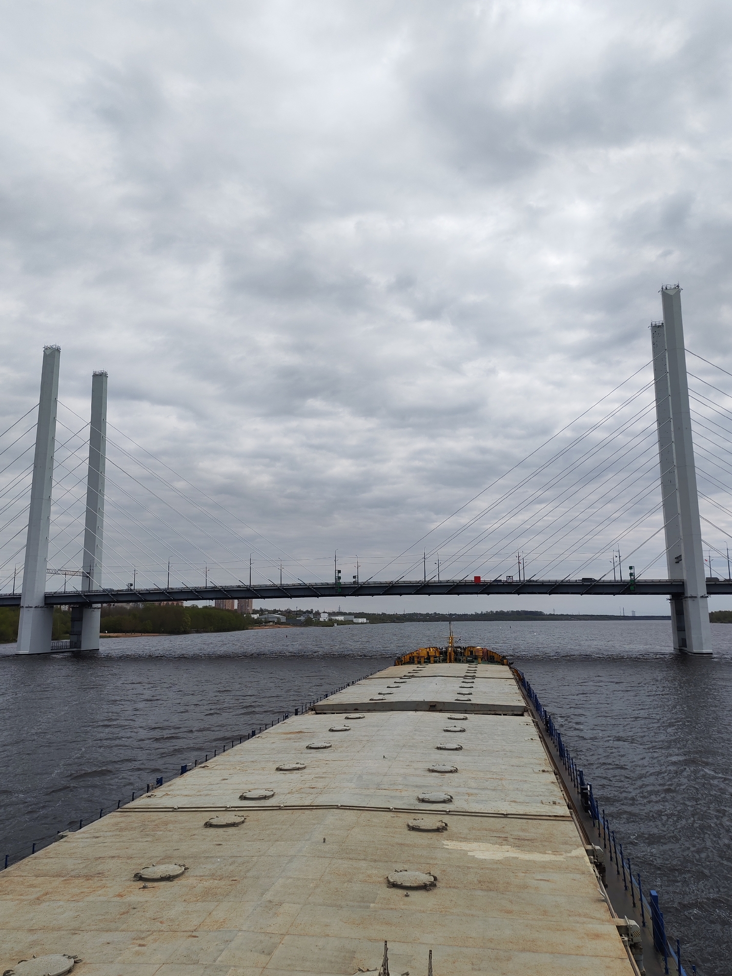 The image shows a wide-angle perspective from the deck of a flat barge or pontoon on the Sheksna River in Cherepovets, Russia. The main subject is the October Bridge, a cable-stayed bridge. The bridge towers are white, and the cables are visible connecting the towers to the bridge deck. The deck of the bridge appears to carry traffic, with some vehicles visible.
The sky is overcast with gray clouds. The water of the Sheksna River is dark and choppy, showing ripples and small waves. The deck of the barge is a dull gray with a pattern of round hatches or access points. Blue railings are visible along the edges of the barge. In the background, beyond the bridge, is a glimpse of the shoreline with trees and some buildings. The overall scene conveys a sense of industrial infrastructure and river transportation in overcast weather.