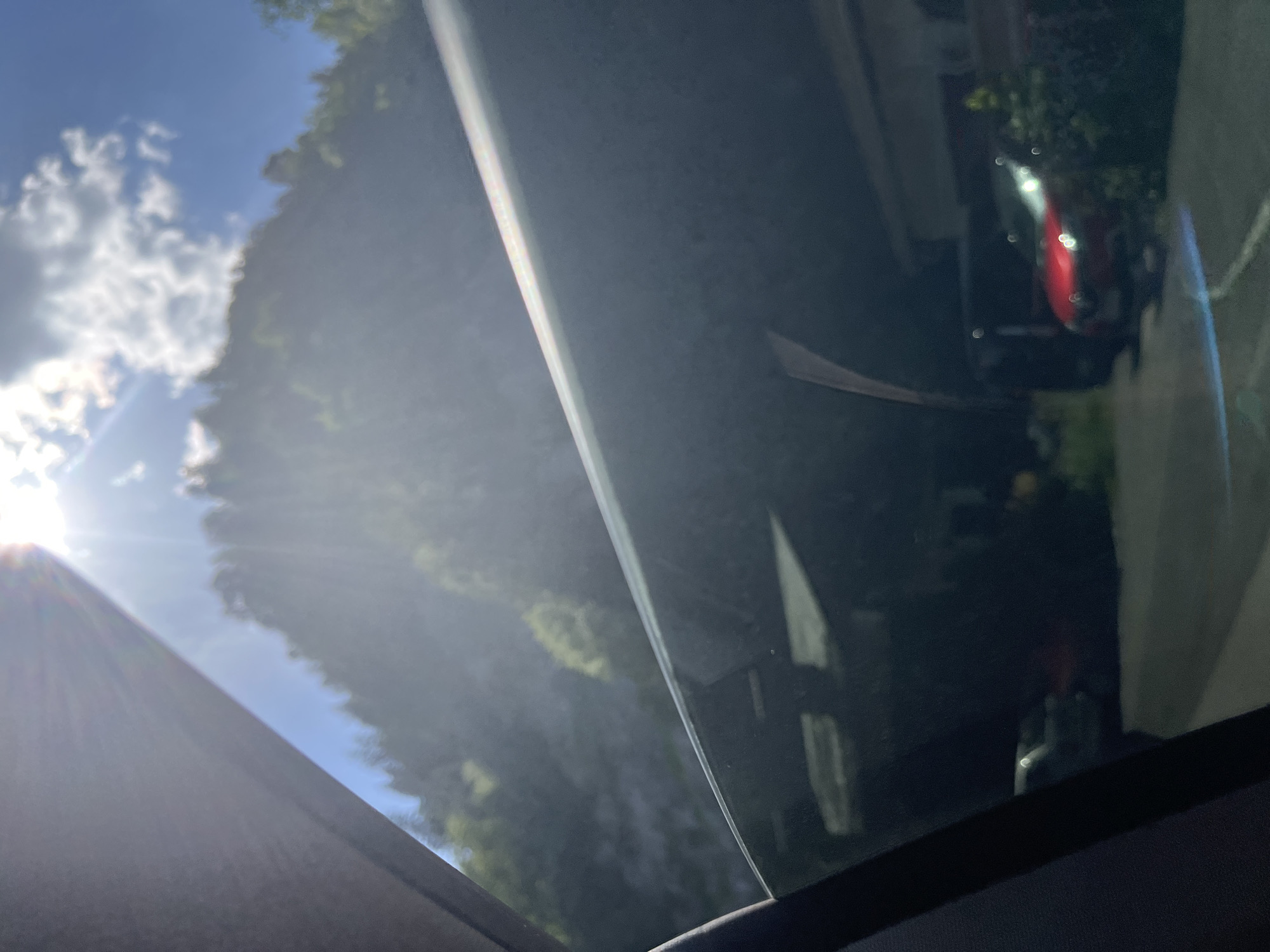 This is a photograph taken from inside a vehicle, looking up through a sunroof. The sun is shining brightly in a blue sky with fluffy white clouds, casting a lens flare across the image. Through the sunroof, one can see trees and foliage, suggesting an outdoor, possibly natural setting. In the upper right portion of the image, there is a reflection in the glass showing a red car parked in what appears to be a driveway or street, with some greenery and part of a building visible. The light suggests it is daytime, likely morning or afternoon given the angle of the sun and the brightness. The overall impression is one of a sunny day, perhaps on a drive or enjoying a clear sky from within a vehicle. No people are visible, and there is no discernible text in the image. The setting appears to be a residential or semi-rural area with trees and a road.