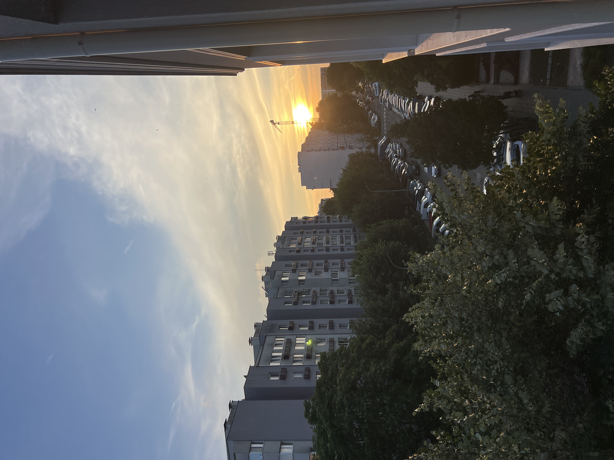 The image captures a sunset scene in the city of Burgas, Bulgaria. The view is from an elevated position, looking down onto a street lined with parked cars and flanked by apartment buildings and trees. The sky is a gradient of blue and orange, with the sun prominently visible near the horizon, partially obscured by a construction crane. Several birds are visible in the sky. The apartment buildings are modern, mid-rise structures with many windows. A large section of a building is visible on the right side of the frame, appearing to be part of a balcony or window ledge. The trees in the foreground are lush and green, with some sunlight filtering through their leaves. The street appears to be a residential area, with a consistent line of cars parked along the side. The overall impression is of a typical urban evening in Burgas, with the natural beauty of the sunset contrasting with the built environment.