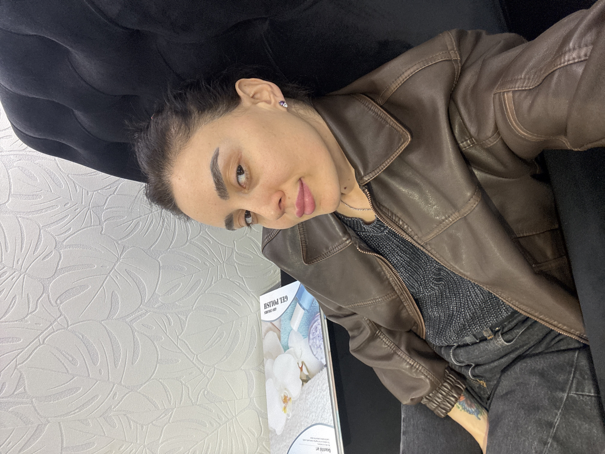 A medium-angle self-portrait shows a person with dark hair and light skin wearing a brown leather jacket and a grey shirt, sitting in a black upholstered chair. The person is looking towards the camera. The background features a white wall with a raised monstera leaf pattern and a promotional sign for gel nail polish. The image appears to have been taken in a salon, possibly in Tbilisi, Georgia, based on the likely service being advertised. No specific details allow the determination of precise location within Tbilisi.