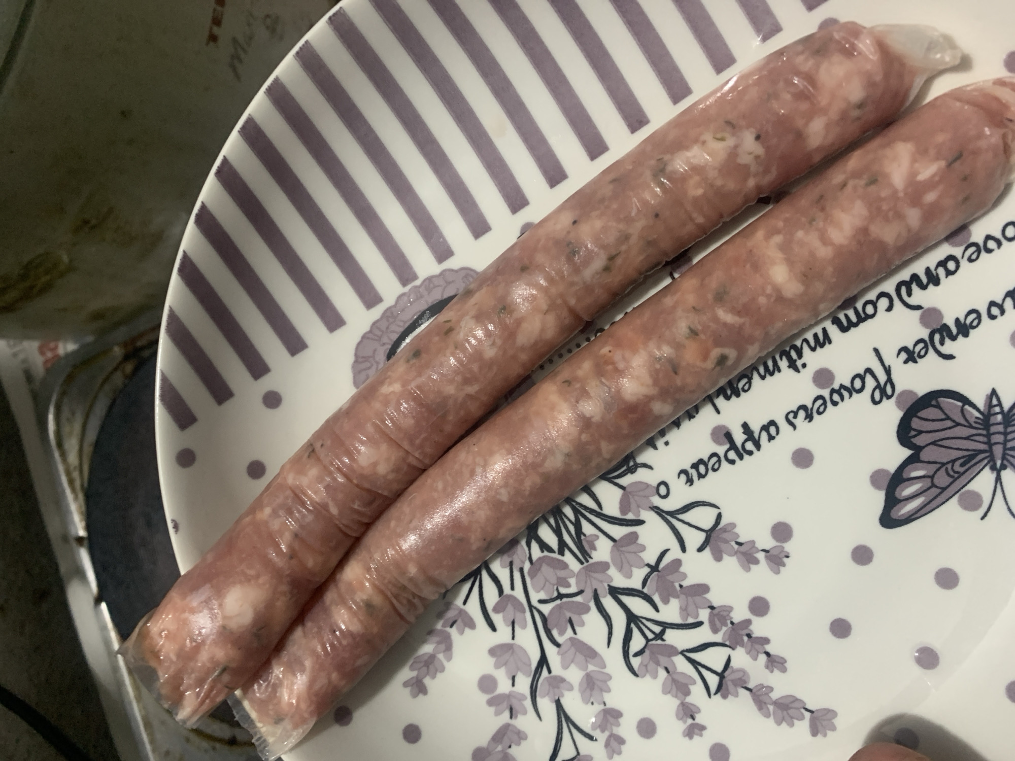 The image shows two raw sausages on a decorative plate. The sausages are elongated and appear to be wrapped in plastic. The plate is white with a pattern of purple stripes and lavender sprigs, along with small purple dots and a butterfly illustration. The sausages are positioned diagonally across the plate. The background is blurred but suggests a stovetop and possibly a pot or pan. The overall scene is likely an indoor setting, possibly a kitchen, given the presence of a stovetop and uncooked food. There is no indication of the city of Mechkarevo, Bulgaria, in the image.