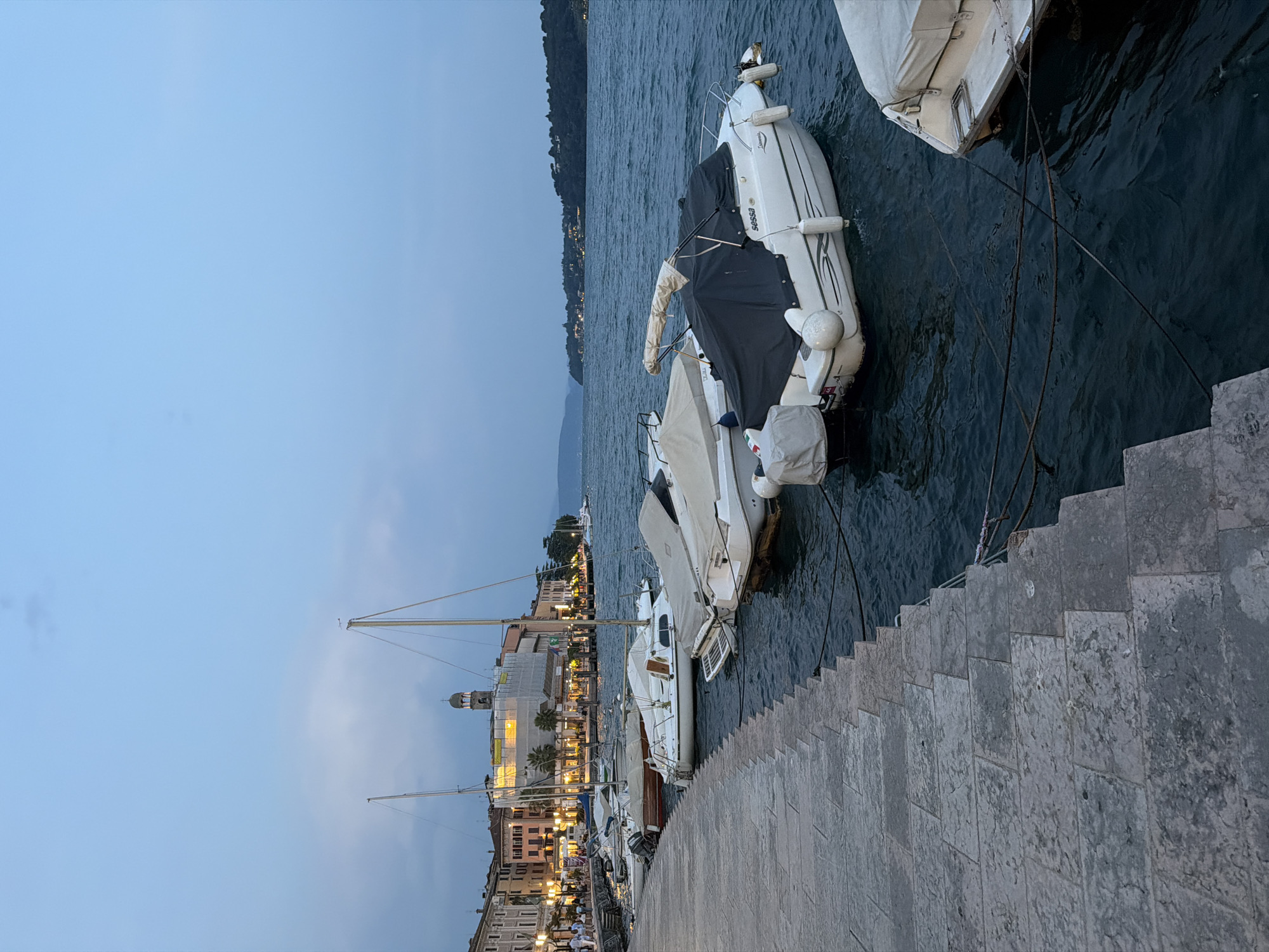 This image depicts a waterfront scene in Salò, Italy, during what appears to be dusk or early evening. In the foreground, a stone walkway lines the edge of the water, with several boats moored alongside. These include small motorboats, some covered with tarps, and at least two sailboats with masts. The boats are bobbing gently on the dark blue water.
Beyond the boats, the shoreline of Salò is visible, characterized by multi-story buildings with illuminated windows, suggesting shops and residences. A prominent church tower with a clock face stands out against the twilight sky, undergoing some sort of construction or renovation, as indicated by scaffolding. Palm trees are interspersed along the waterfront promenade, adding a touch of greenery. In the distance, rolling hills and the expanse of the lake can be seen, fading into the hazy horizon. The sky is a pale blue, transitioning from a lighter shade at the top to a darker hue closer to the horizon, with a few wisps of clouds. The overall atmosphere is serene, with the lights from the town reflecting faintly on the water.