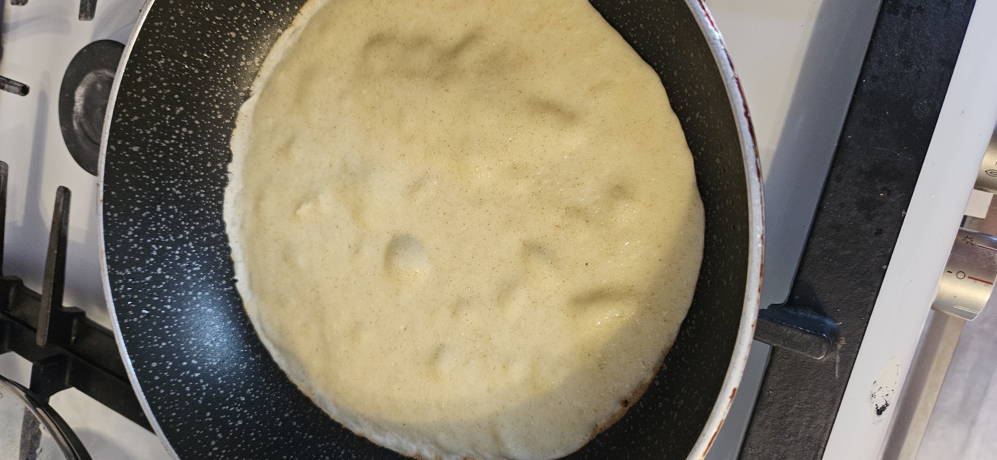 An overhead, close-up shot shows a pancake cooking in a non-stick pan on a white gas stove. The pancake is light brown and has several small bubbles on its surface. The pan has a black speckled interior and a red and white exterior rim. The stove has black grates and white burner covers. The image does not provide enough information to determine the location in Vitsebsk, Belarus.