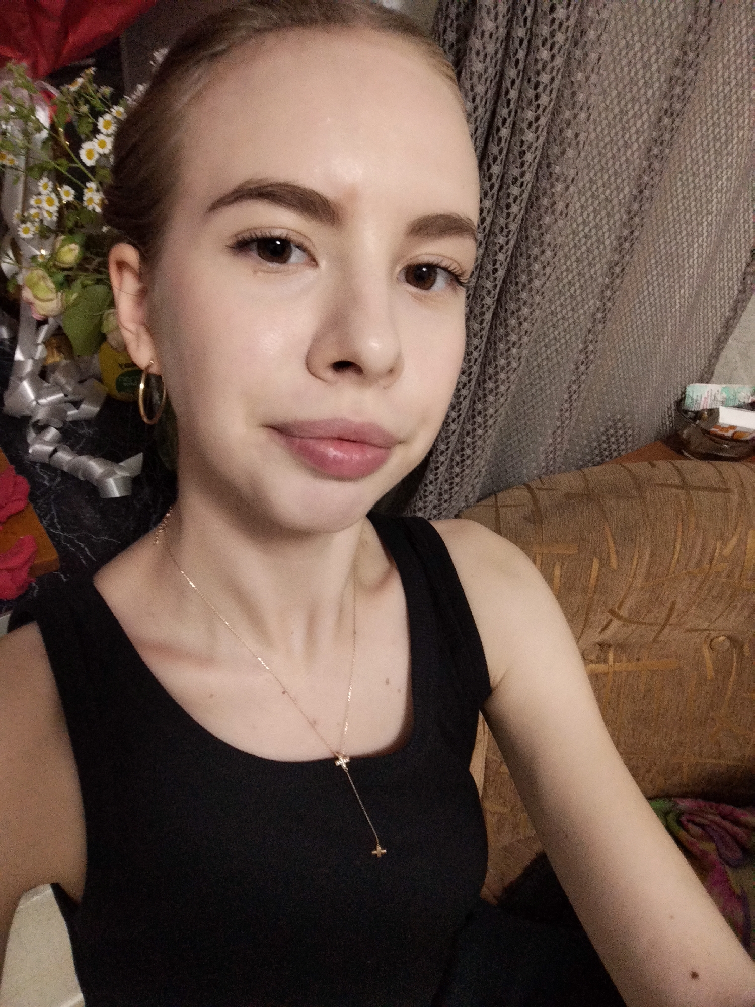 A young Caucasian woman with light skin and brown hair is looking at the camera in a medium shot. She is wearing a black tank top and a gold necklace with a small cross pendant. She has a hoop earring in her left ear. Her makeup appears to be natural. In the background, there is a curtain with a knitted pattern and a couch. To the left of the woman, there are some flowers and gift wrapping. The scene appears to be indoors.