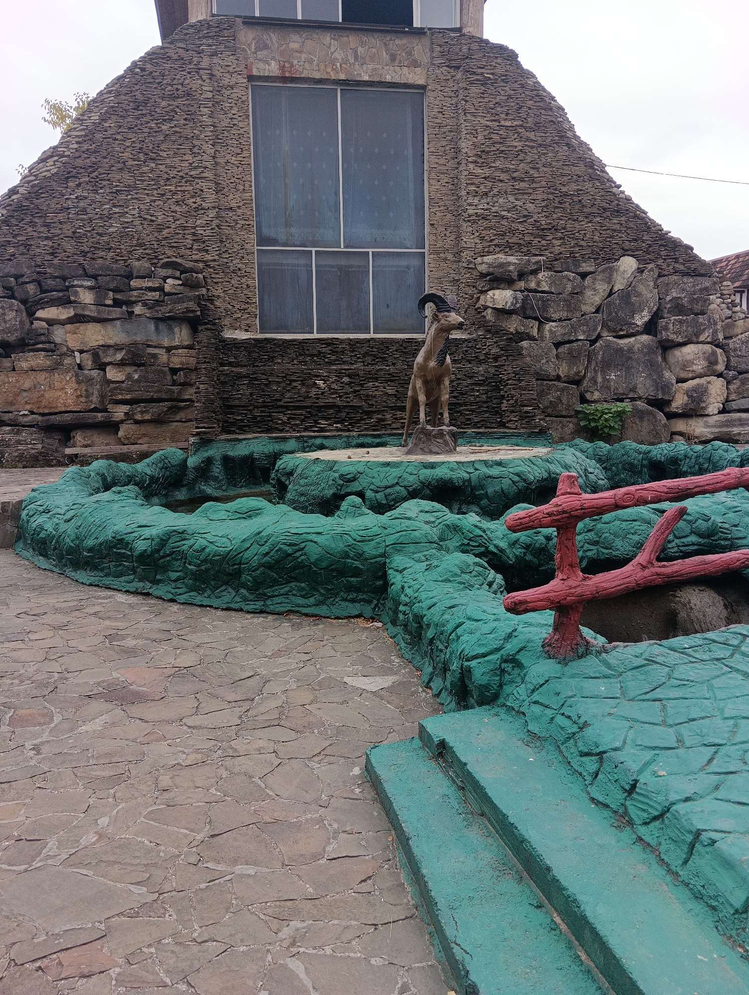 A statue of a goat stands in front of a building with a stone facade and a large window. The statue is positioned within a green, textured, pond-like structure. The ground is paved with irregular stones. A red, branch-like railing is visible in the foreground, and there are steps leading up to the green structure. The sky is overcast.