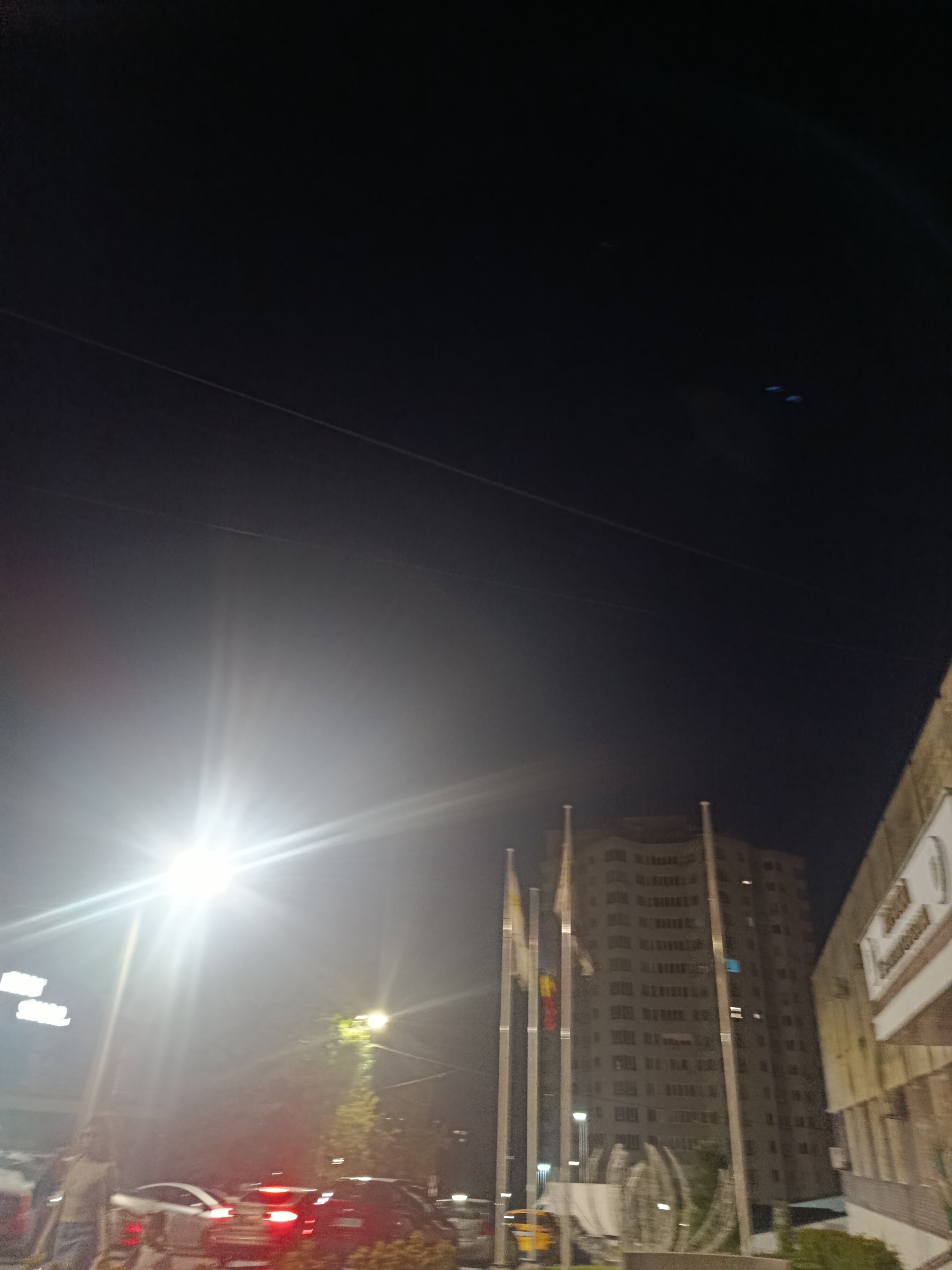 The image depicts a nighttime scene in Chișinău, Moldova, with a prominent street light on the left side casting a bright glare that overexposes a portion of the foreground. In the middle ground, a tall, modern building with many windows is visible, flanked by flagpoles. The flags on the poles are partially obscured but appear to have red, yellow, and blue colors. In the foreground, several cars are visible, some with their brake lights illuminated, suggesting they are stopped or moving slowly in traffic. The environment appears to be a city street, with some greenery and pedestrian areas visible. The sky is dark, indicating it is late at night.