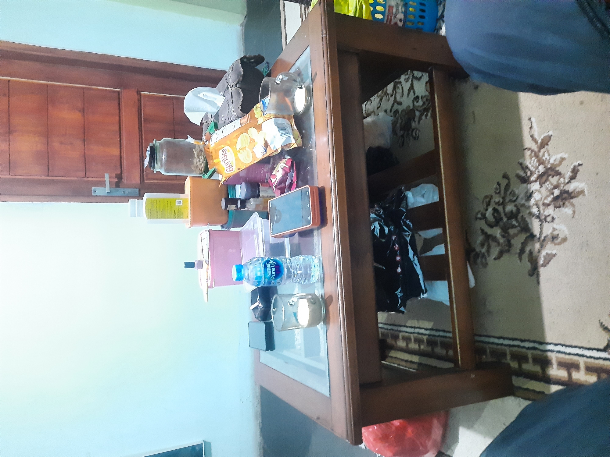 In a room, likely a living room, in Bandung City, Indonesia, a wooden coffee table with a glass top is laden with various items. Positioned on the table are a bottle of Nestle Pure Life water, a glass containing a light-colored liquid, a cell phone, an orange-cased smartphone, a pack of GuriBee crackers, a glass jar filled with small objects, a roll of white paper towels, a small pink container, and a few other bottles and containers. A wooden door with horizontal panels is visible in the background. The floor is covered with a patterned rug, and to the left of the table, a red plastic bag is present. The overall scene suggests a casual, everyday setting.