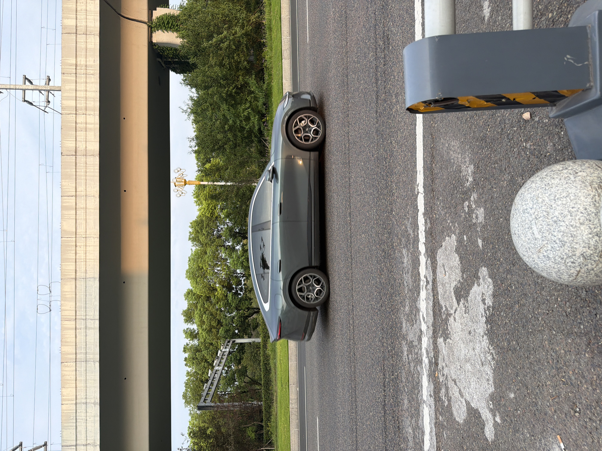 A gray sedan is driving on a paved road. The car has tinted windows and a sleek, modern design. In the foreground, there is a granite sphere and a traffic barrier. In the background, there are trees and an elevated highway structure with power lines. The scene appears to be in Jiangjin, China, based on the provided context.
