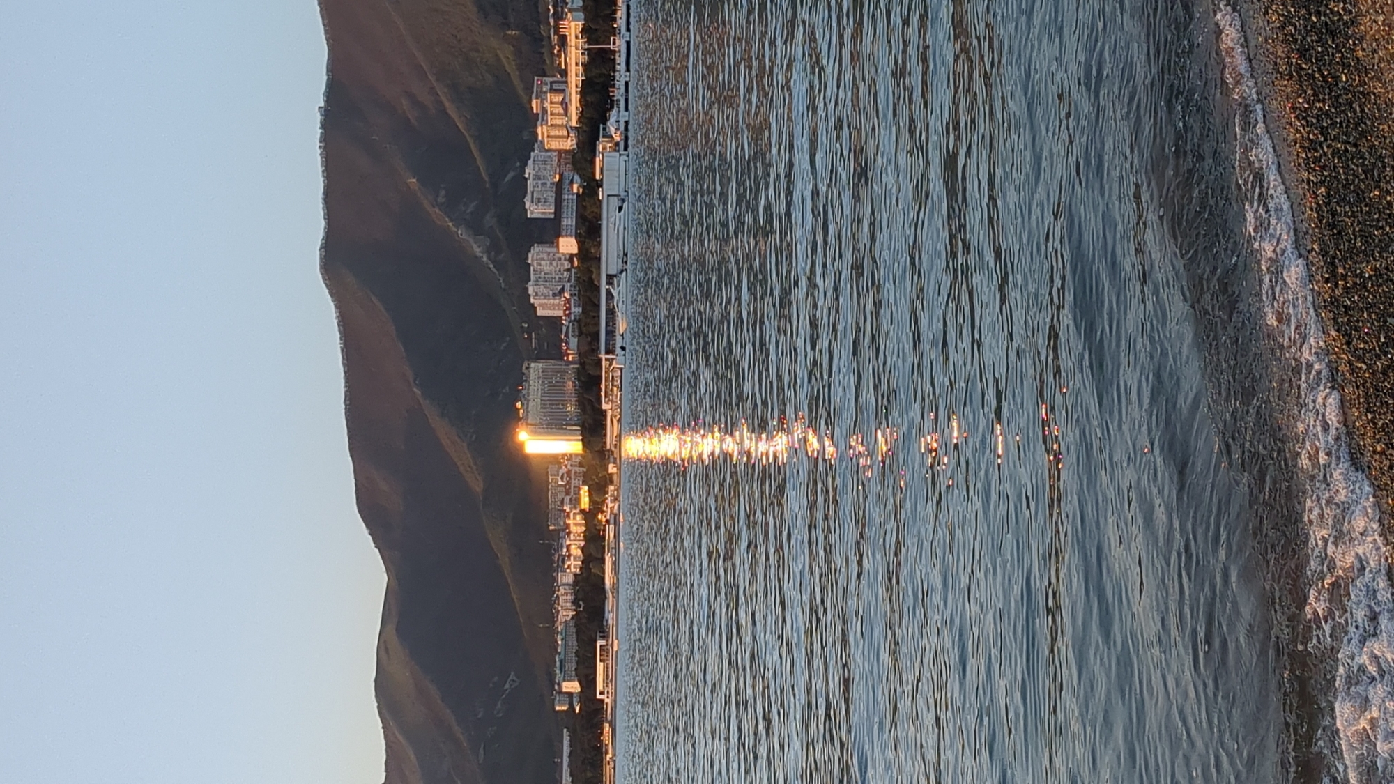 The image displays a tranquil coastal scene, likely at sunrise or sunset, given the warm, golden light and long reflections on the water. The perspective is oriented with a clear sky occupying the top portion, transitioning to dark, vegetated mountains or hills that dominate the left side of the frame. Along the base of these mountains, an urban development of buildings, possibly hotels or residential structures, lines the shoreline. The sun's rays catch the facades of some buildings, causing them to glow with a bright, golden light.
The central and right-hand side of the image is dominated by a large body of water, possibly a sea or a substantial lake. The water's surface is rippled with small waves, and a prominent streak of shimmering golden light, reflecting the low sun, extends from near the buildings towards the foreground. To the far right, a narrow strip of coarse-grained beach, appearing to be made of pebbles or dark sand, is visible, with gentle white-edged waves lapping at its edge. The overall atmosphere is serene and peaceful. There are no people or discernible text in the image.