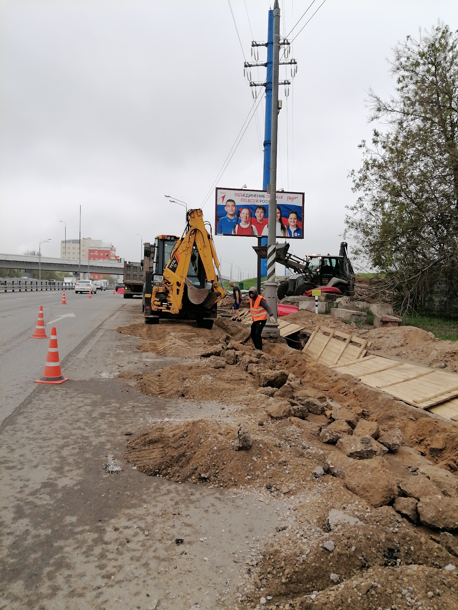 The image depicts road construction along a roadway in Moscow, Russia. A yellow backhoe is parked on the shoulder of the road, next to a line of orange traffic cones. A worker in a reflective vest stands beside an excavated trench lined with wooden planks. Another backhoe is visible behind the trench. In the background, cars drive along the roadway, passing under an overpass. A building is visible in the distance. A large billboard with images of children on it is positioned above the roadwork. Overhead power lines are visible. The sky is overcast.