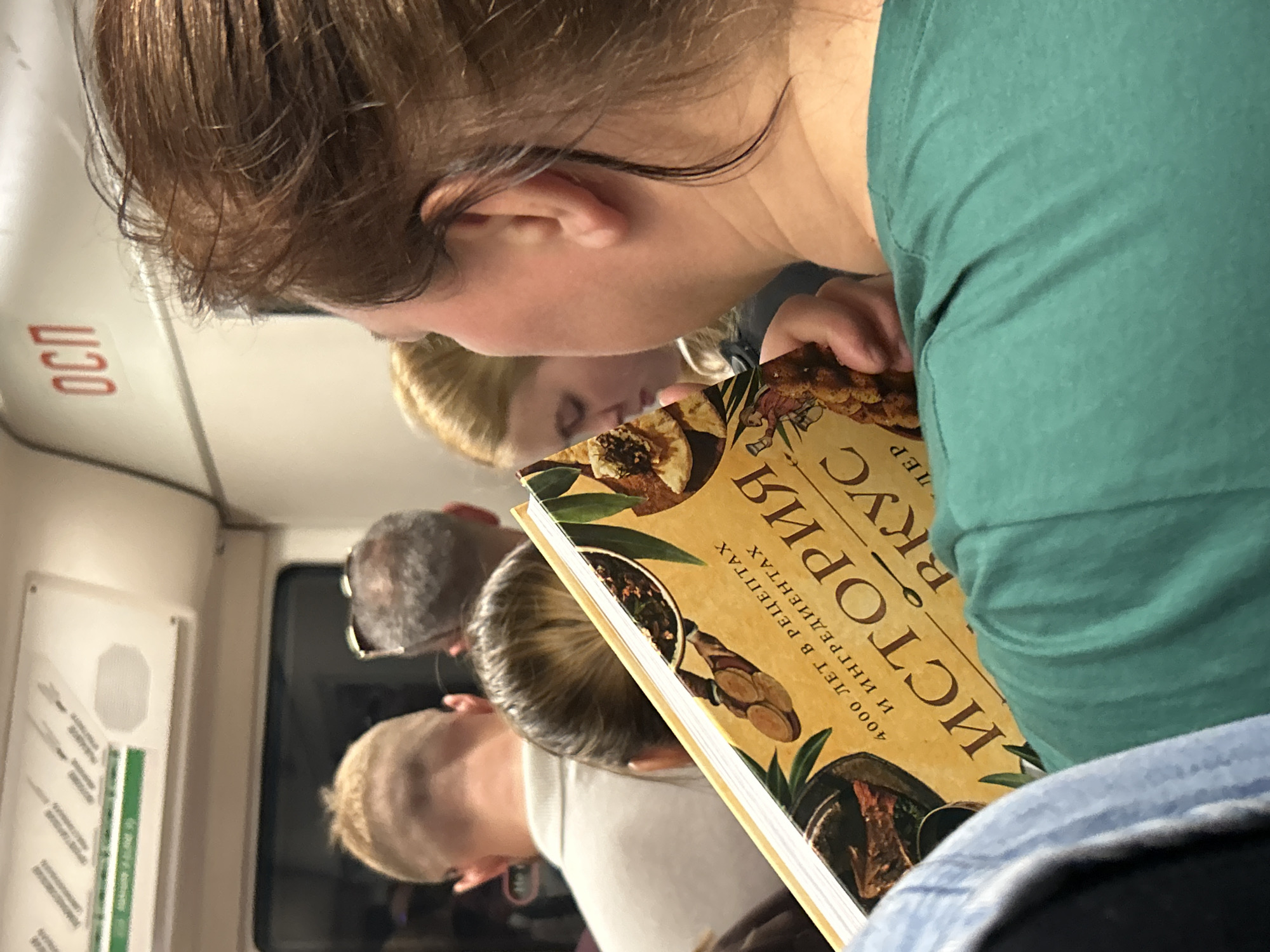 In this close-up, medium-shot photograph, a woman with brown hair is seen from the side, wearing a green t-shirt. She is holding a yellow book open with both hands, her right hand is visible, and she is looking down at it. The book has text in Cyrillic, with prominent letters "ИСТОРИЯ" and "ВКУС." Above the text, there appears to be an illustration featuring food items. In the background, other people are visible, including a person with blond hair looking down, and individuals with short haircuts in the distance. The environment appears to be the interior of a public transport vehicle, possibly a metro train, indicated by the overhead lighting, signage with Cyrillic text ("ОСП"), and seating arrangements implied by the composition. The overall lighting suggests an indoor, likely artificial light setting, common in St. Petersburg's metro system.