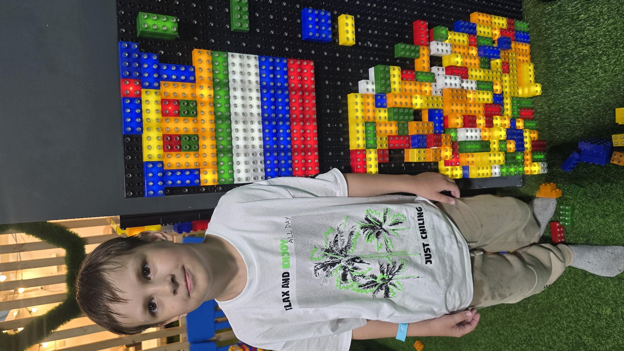 The image depicts an indoor recreational area, possibly located in Izhevsk, Russia, with a young male child standing beside a large, illuminated building block display.
The central figure is a boy, estimated to be between 8 and 10 years old, with short brown hair and dark eyes, looking directly at the camera with a subtle smile. He is wearing a white short-sleeved t-shirt with a graphic design that includes the text "RELAX AND ENJOY ALL DAY" above a grayscale image of three palm trees outlined in neon green, and "JUST CHILING" below. He is dressed in light brown or khaki-colored sweatpants and light grey socks. A light blue wristband is visible on his left wrist.
To the boy's right, a prominent black pegboard display is covered with numerous translucent building blocks that emit light. The upper section of the display features a horizontal pattern resembling the Russian flag, composed of distinct rows of white (or clear), blue, and red illuminated blocks. Below this, and extending downwards, a complex, multi-colored structure of various light-up blocks in yellow, red, blue, green, white, and orange forms an abstract design. Several loose, colorful building blocks are scattered on the green artificial grass floor near the base of the display.
The floor of the scene is covered with bright green artificial turf. The background, out of focus, reveals a wooden slatted structure with warm yellow string lights visible through it. In the upper left corner, a decorative element of green, possibly artificial, foliage is present. A blue child-sized chair is partially visible in the far left background. The overall lighting is bright, indicative of an indoor entertainment or play facility.