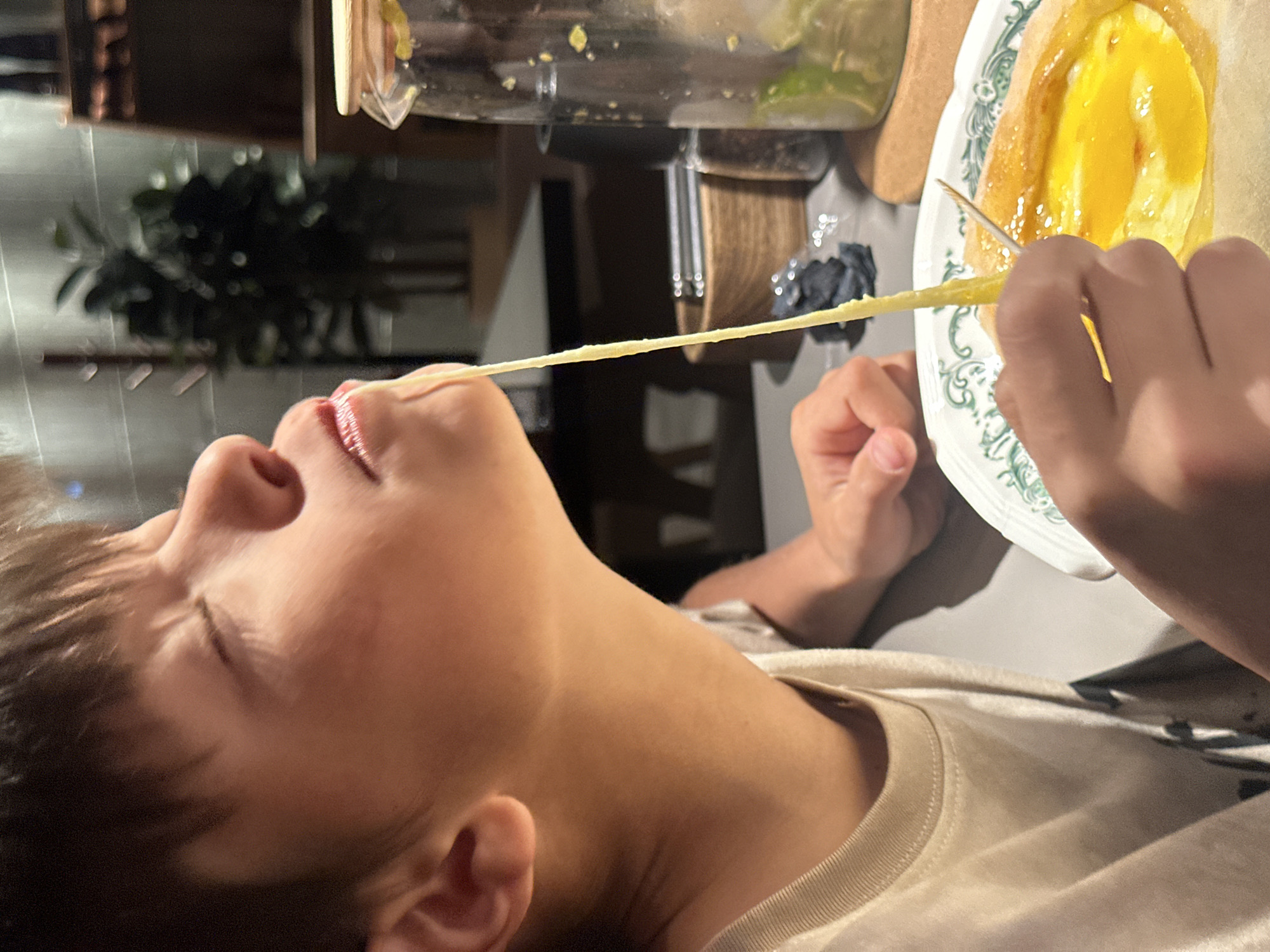 A child with dark hair is lying on their back with their eyes closed and mouth open. A strand of yellow food stretches from a plate, held by the child's hand, to the child's mouth. The plate appears to contain a cooked dish with a yellow topping. The child is wearing a light-colored shirt. In the background, there is a jar, a table, and some blurred objects. There is no evidence to connect this image to Chaikovsky, Russia.