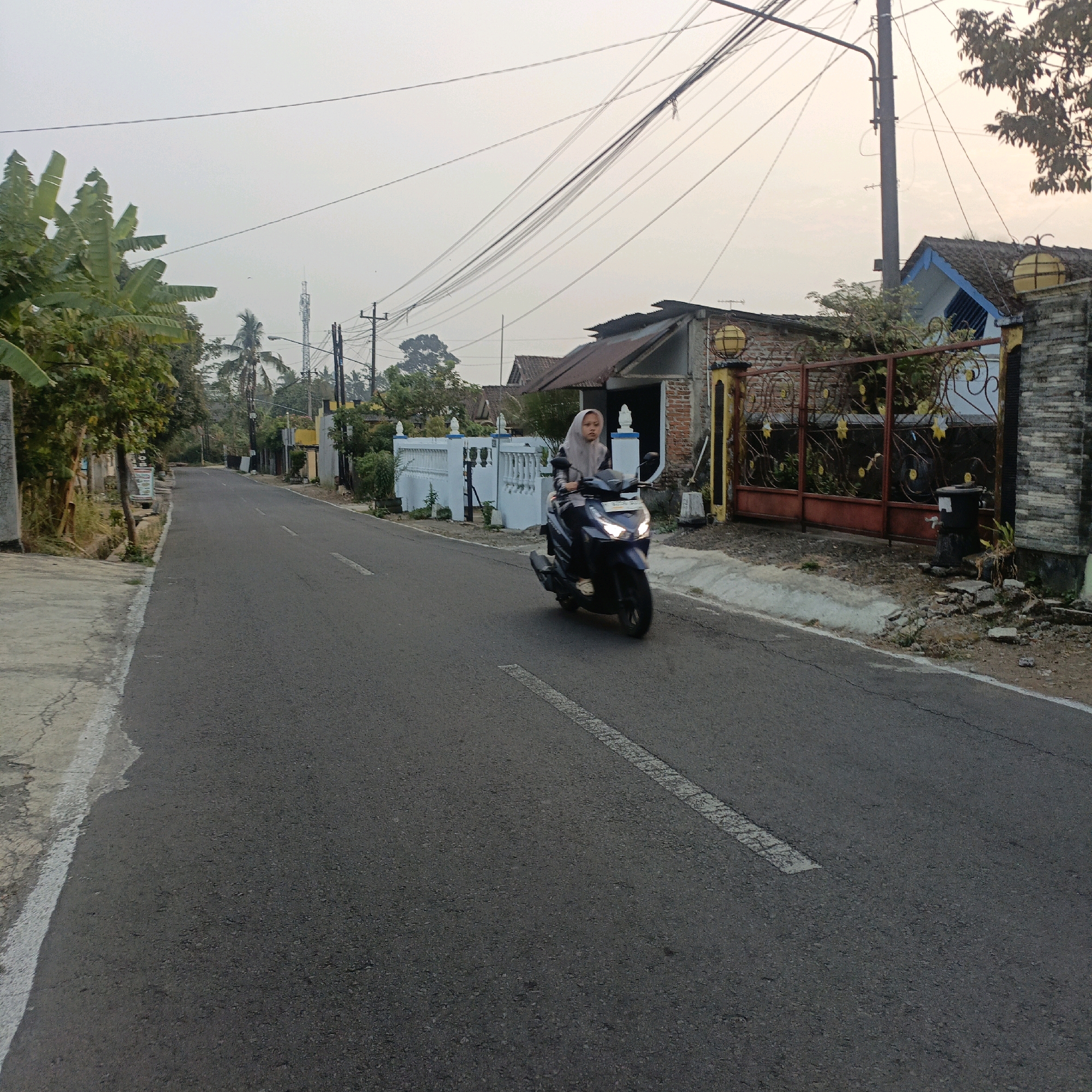 A person is riding a dark blue motorcycle on a paved road in an outdoor urban environment. The person is wearing a light grey hijab and a dark jacket. The motorcycle has its headlights on. The road is asphalt with a broken white line marking the center. On the right side of the road, there is a brick house with a dark-colored roof and a decorative gate. The gate is red with yellow swirling metalwork. Behind the gate, there are trees and vegetation. There are also utility poles and wires overhead. On the left side of the road, there is a concrete sidewalk, vegetation, and more houses in the background. The sky is overcast and appears to be either dawn or dusk. The image appears to have been taken in Salatiga, Indonesia, given the presence of a cell tower in the background.