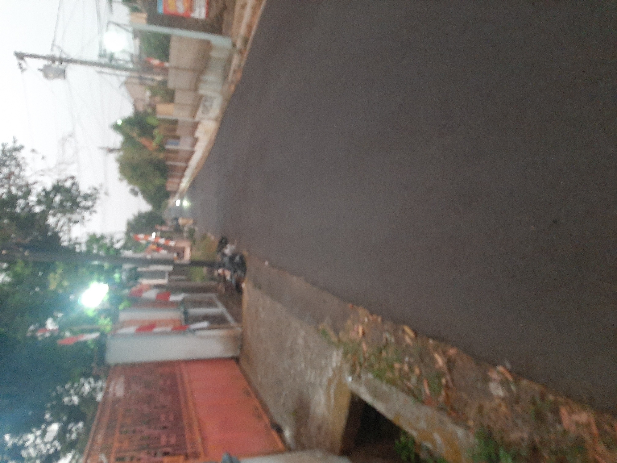 The image depicts a street scene in Purwanegara, Indonesia, likely taken in the late afternoon or early evening. A paved road occupies the right side of the frame, stretching into the distance. On the left side of the road, there is a reddish-orange fence with decorative metalwork. Adjacent to the fence is a concrete walkway that runs alongside the road, with a drainage ditch visible at the bottom left. Several Indonesian flags, red and white, are mounted on poles along the side of the road, some appearing to be attached to a white structure. Trees with green foliage are present, casting shadows and contributing to the slightly dim lighting. Electrical poles and wires are visible in the background, along with some buildings. A few blurry figures and what appear to be trash bags are present on the side of the road near the flags. The overall atmosphere is somewhat muted, with a soft focus that suggests a low-light condition or a less-than-ideal camera.