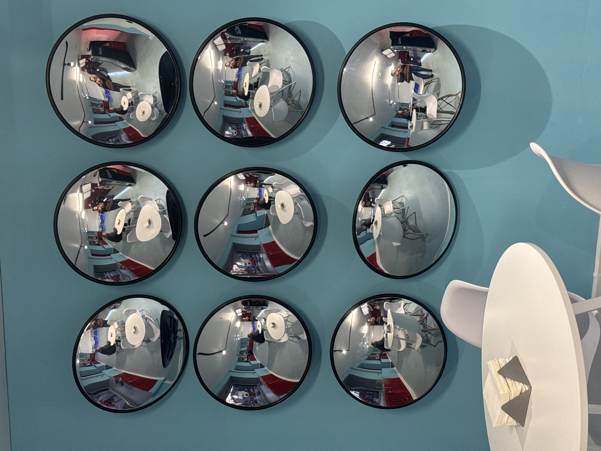 The image shows nine convex mirrors arranged in a 3x3 grid on a blue wall. The mirrors reflect a distorted view of a room with chairs, tables, and people. Some mirrors show a man and a woman standing and talking in the room. The reflections suggest the room has blue and red accents. Adjacent to the mirrors on the right side of the image is a white chair with a napkin holder. The environment appears to be indoors. Based on the metadata, the location is in Chaikovsky, Russia.