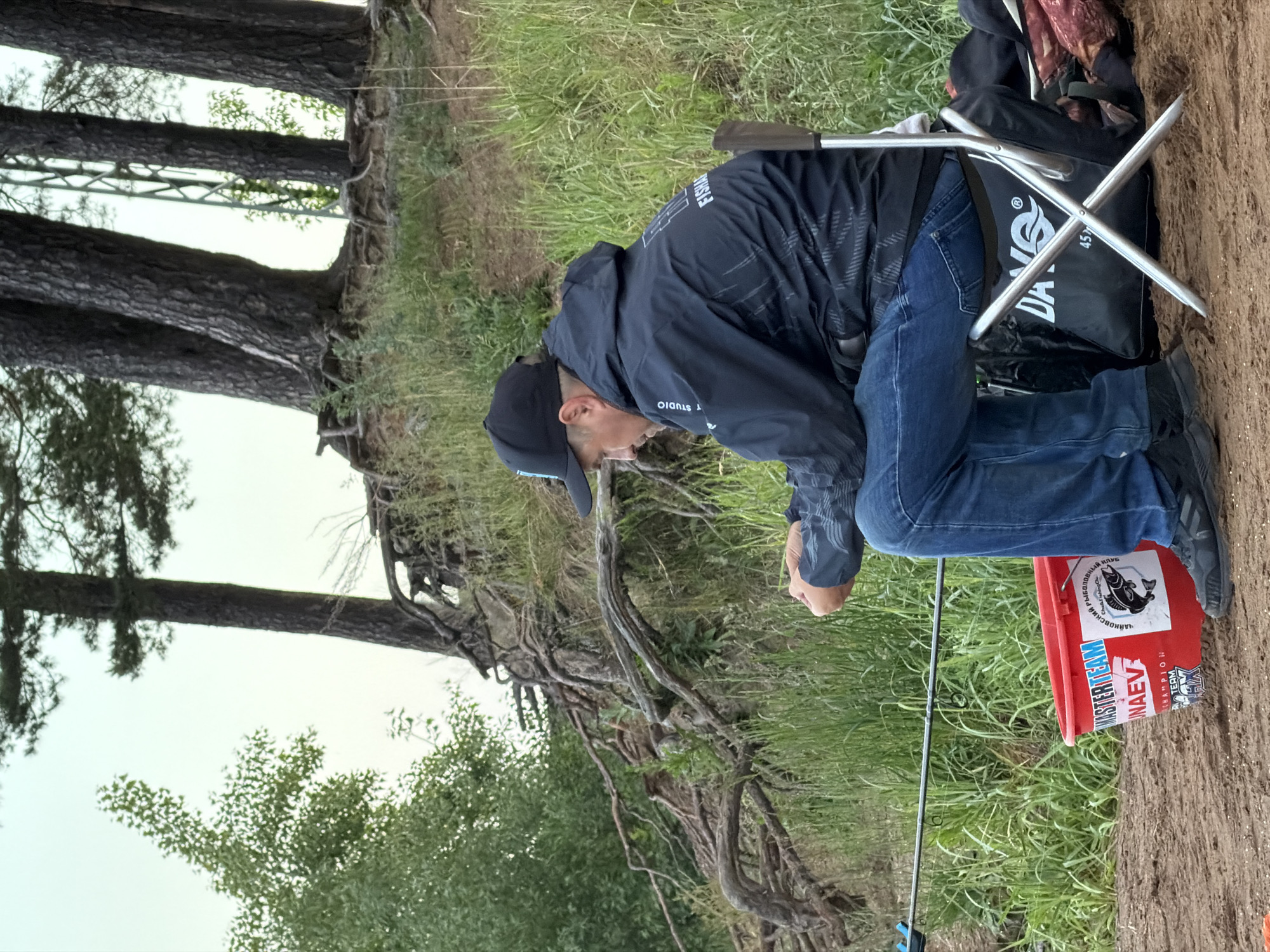 The image displays an outdoor scene, likely a river or lake bank in Chaikovsky, Russia. A person is prominently featured, seated on a silver-colored portable folding chair. They are dressed in a dark blue or black hooded jacket, blue jeans, and a dark baseball cap, leaning forward with their gaze directed downwards, seemingly focused on their hands or fishing equipment.
A fishing rod is visible extending from the lower right portion of the frame towards the left. Near the person's feet is a red plastic bucket adorned with multiple stickers. Visible text on the bucket includes "Чайковский рыболовный клуб" (Chaikovsky Fishing Club) and "СЛАЙМ" in Cyrillic, alongside English text such as "MASTERTEAM", "NAEV", and "CHAMPION TEAM FX". Behind the person, a black bag with a "DAIWA" logo and the number "45" is resting on the chair's back, next to another patterned bag.
The background on the left features several large trees with extensive, exposed roots, suggesting a well-established natural environment. Beyond the immediate sandy ground, there is an area of vibrant green grass. In the distance, between the tree trunks, a faint metal structure, possibly a bridge or support tower, can be discerned against a light, possibly overcast, daytime sky. The scene conveys a sense of calm and focused engagement in fishing.