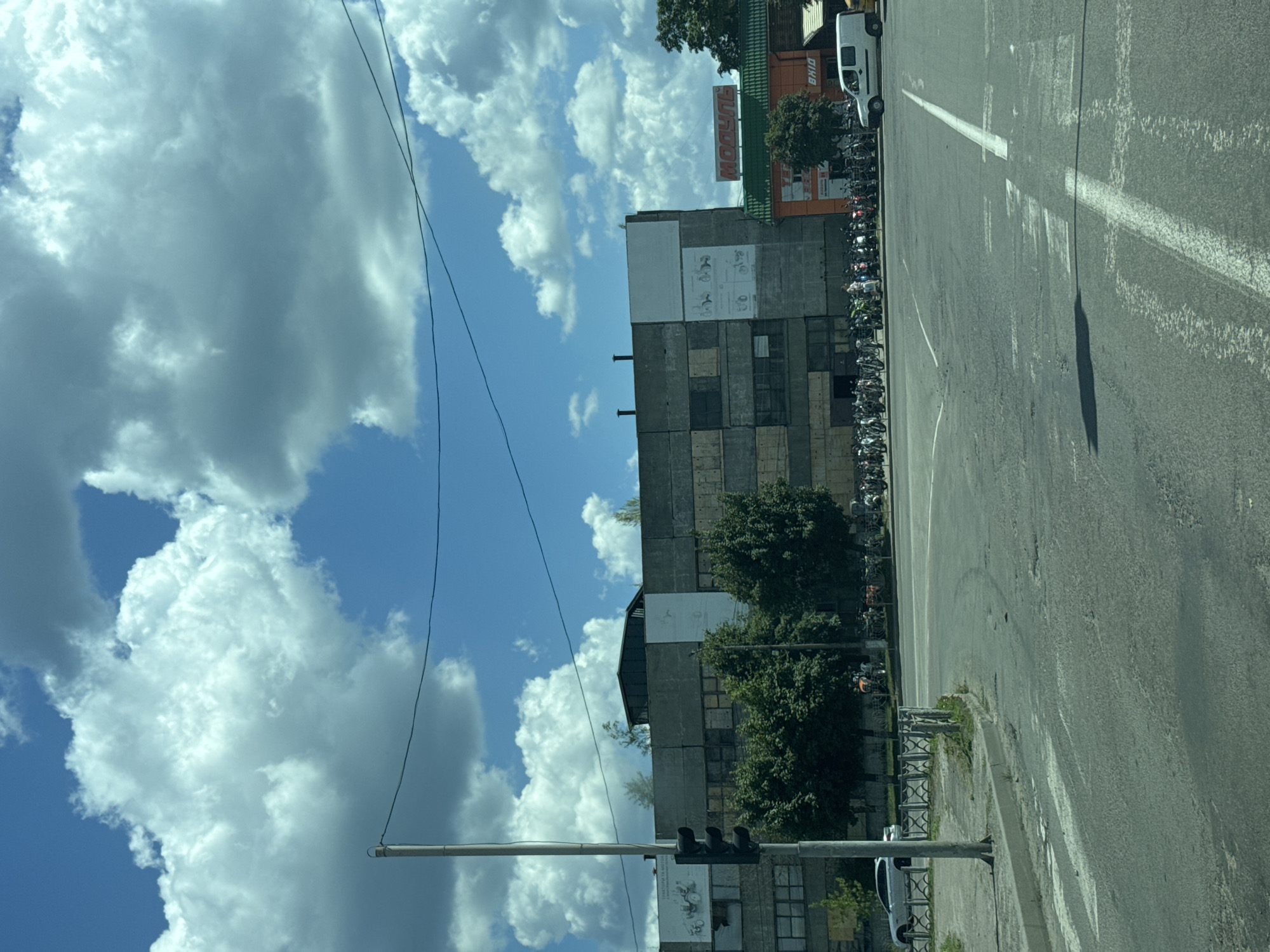 This image depicts an outdoor scene with a large, weathered industrial-style building on the right, juxtaposed against a bright blue sky filled with fluffy white clouds. The building appears to be made of concrete panels and has numerous windows, some of which are boarded up. Trees are visible growing near and on top of the structure. In front of the building, a long row of bicycles is parked, suggesting a public space or facility. A white van is parked further down the street, near the bicycles.
The foreground shows a section of an asphalt road with white road markings, indicating a street or parking lot. A traffic light is visible on a pole in the lower left quadrant of the image. The orientation of the image is rotated, with the sky occupying the left half and the building and road on the right.
There is visible text on a sign on the building that reads "BHID". Another sign with red lettering on an orange background is also visible higher up on the building, but the text is partially obscured and difficult to read definitively. A poster or mural with diagrams of figures is on the side of the building.
The lighting suggests it is daytime, likely late morning or afternoon, given the brightness of the sky and the shadows cast on the road. The weather appears to be fair and sunny, with scattered clouds. The overall impression is one of an urban or industrial setting in Sarny, Ukraine, on a clear day. There are no people prominently featured in the image.